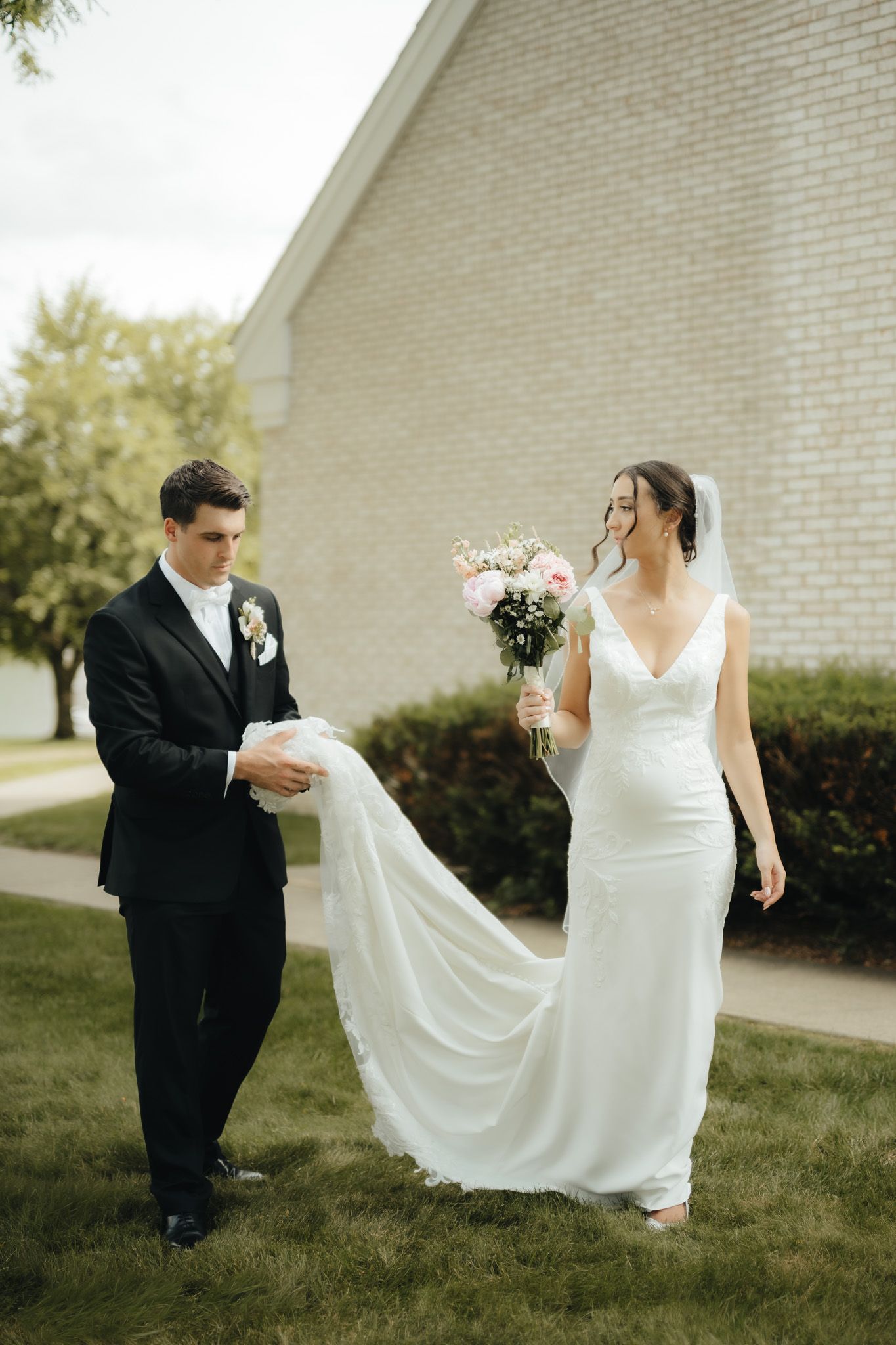 Bride in white gown holding floral bouquet walks with groom in black suit carrying her train outside a brick church.