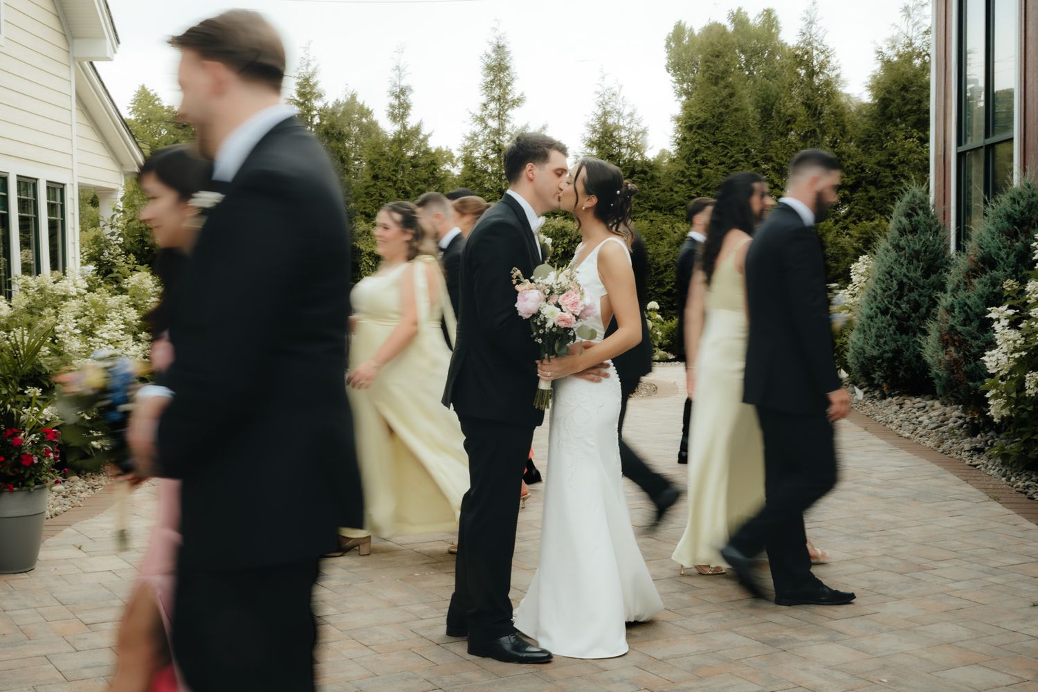 A bride and groom share a kiss surrounded by bridesmaids in yellow dresses and groomsmen in tuxedos outdoors.