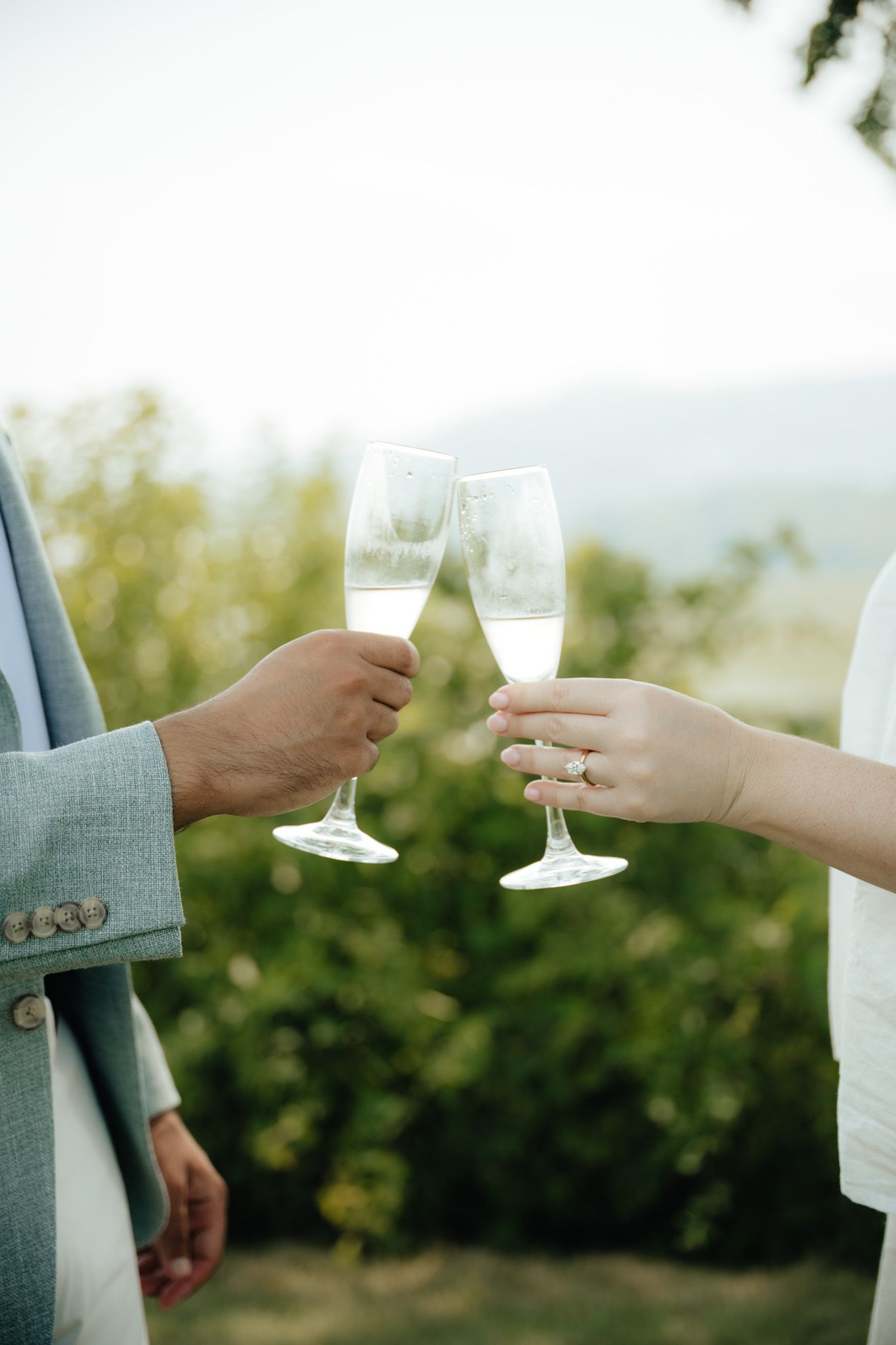 Two hands clinking champagne flutes outdoors, with a diamond engagement ring visible on the bride's hand.