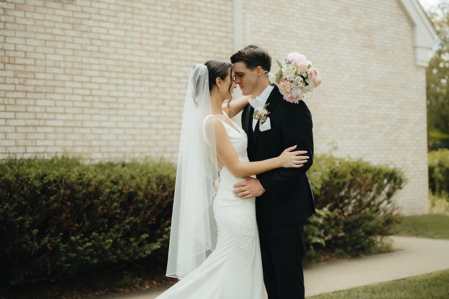 Bride in white gown and veil holds bouquet while embracing groom in black suit outside brick church.