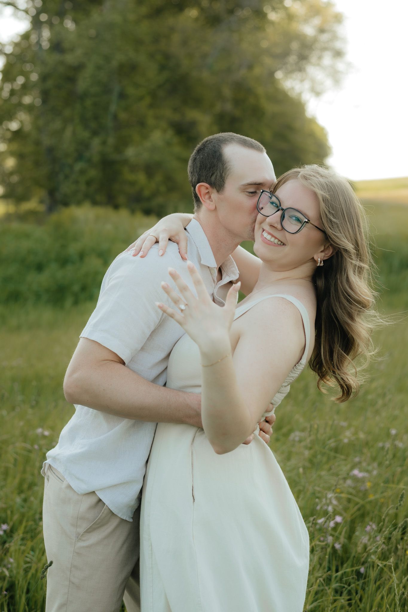 Couple embracing in a sunny green field, woman smiling with engagement ring visible, man kissing her cheek.