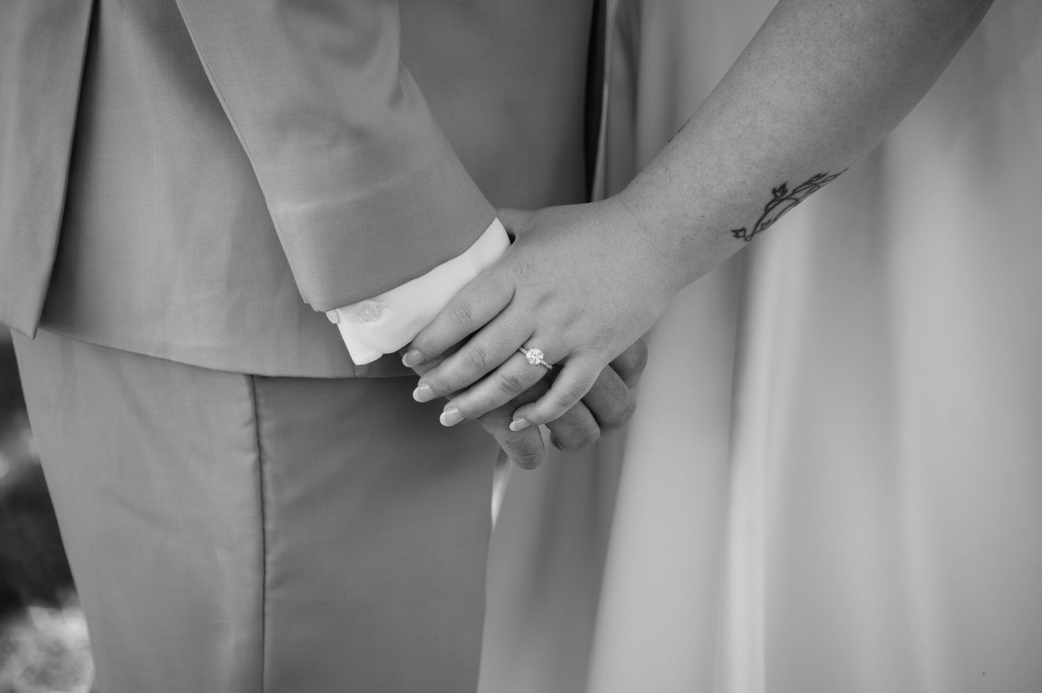 Black and white close-up of bride and groom holding hands, showing wedding ring and formal attire.