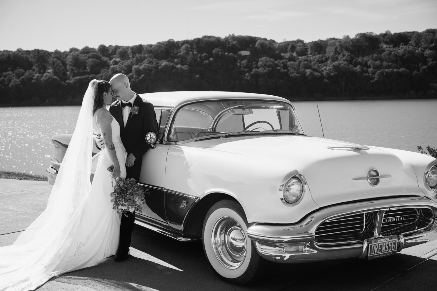 Bride and groom kissing beside vintage classic car near waterfront in black and white photo.