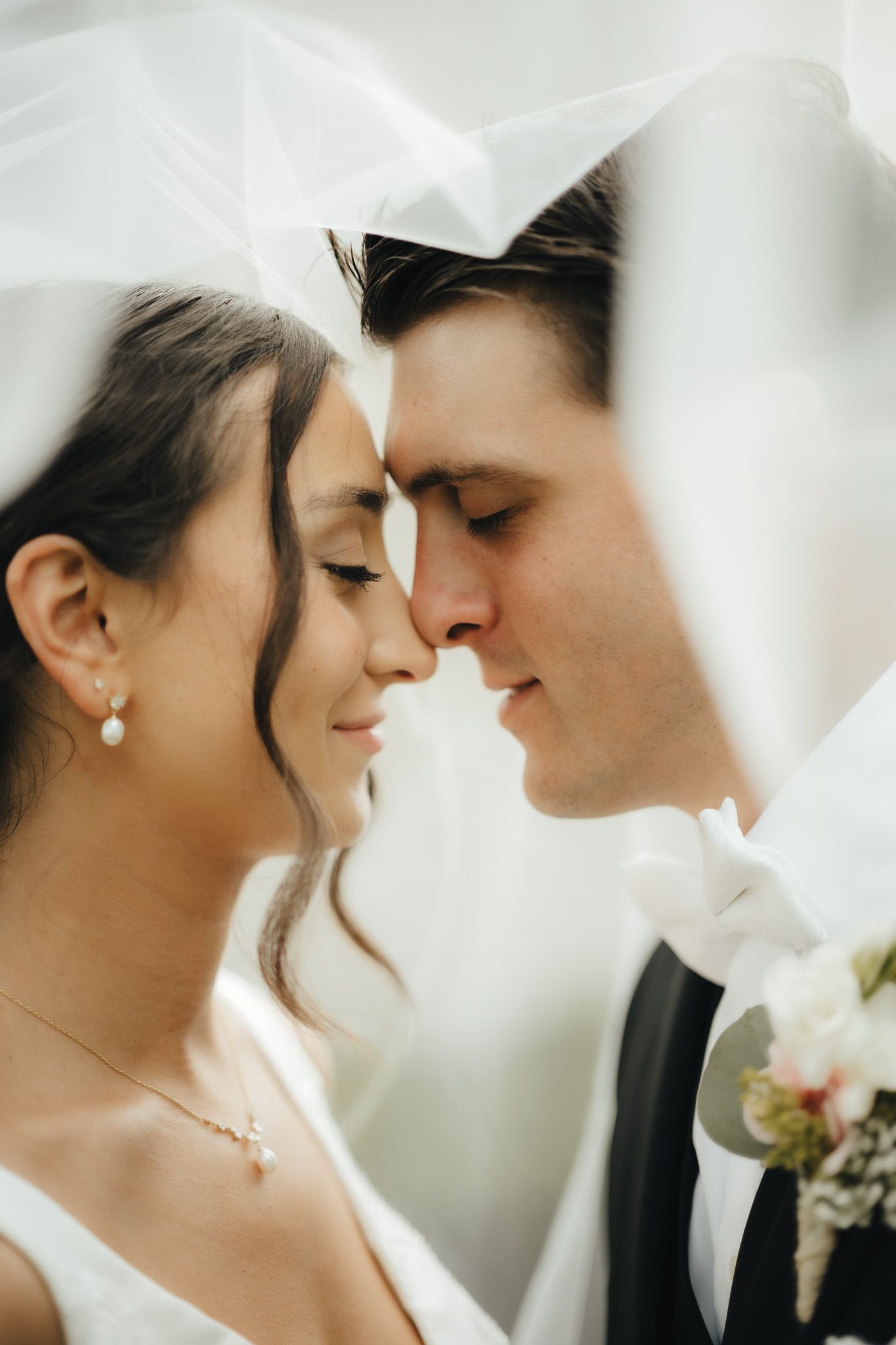 Bride and groom share an intimate moment under a white veil, foreheads touching, eyes closed on their wedding day.