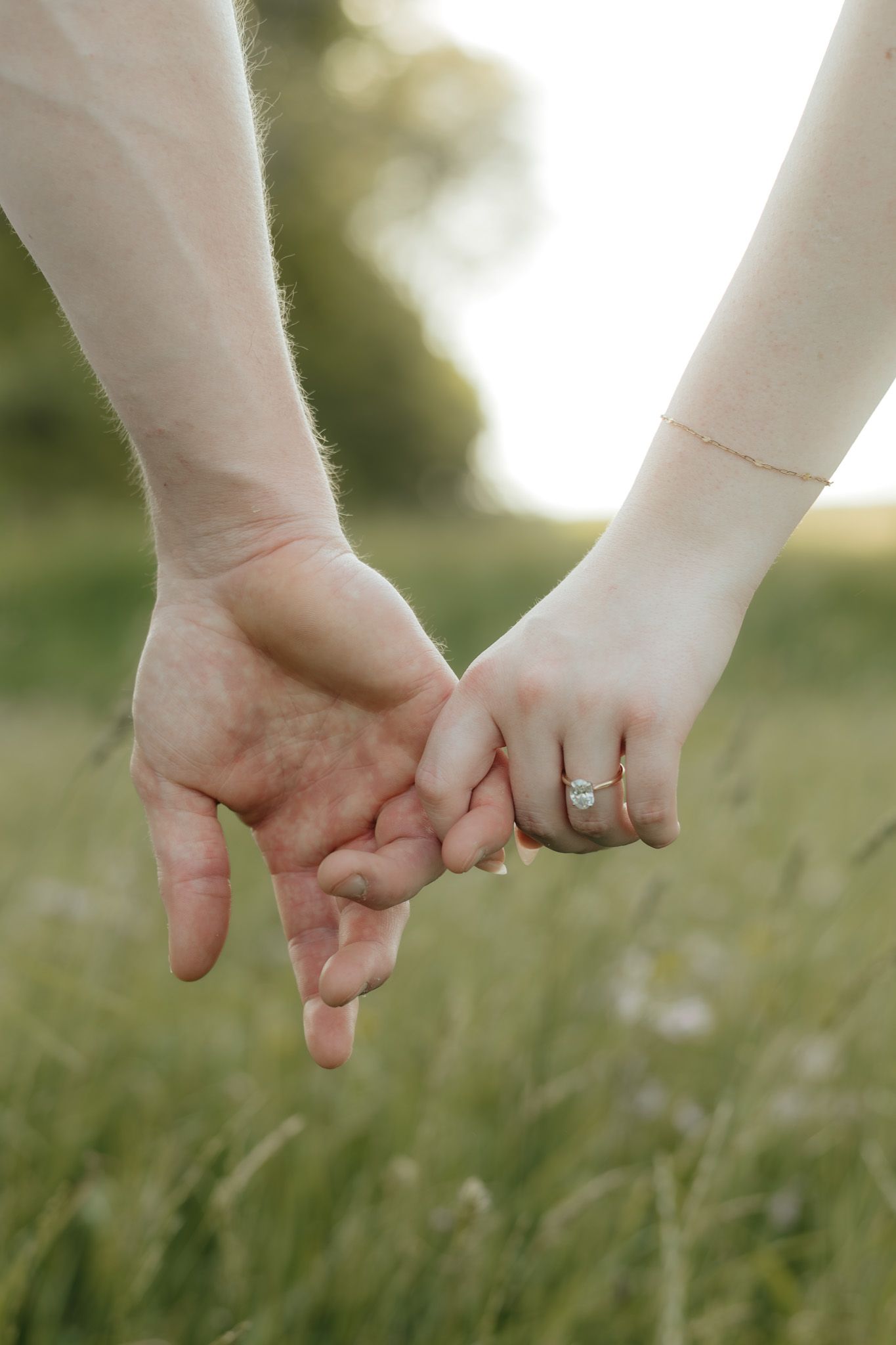 Couple holding hands in a green field, woman wearing a diamond engagement ring and gold bracelet.