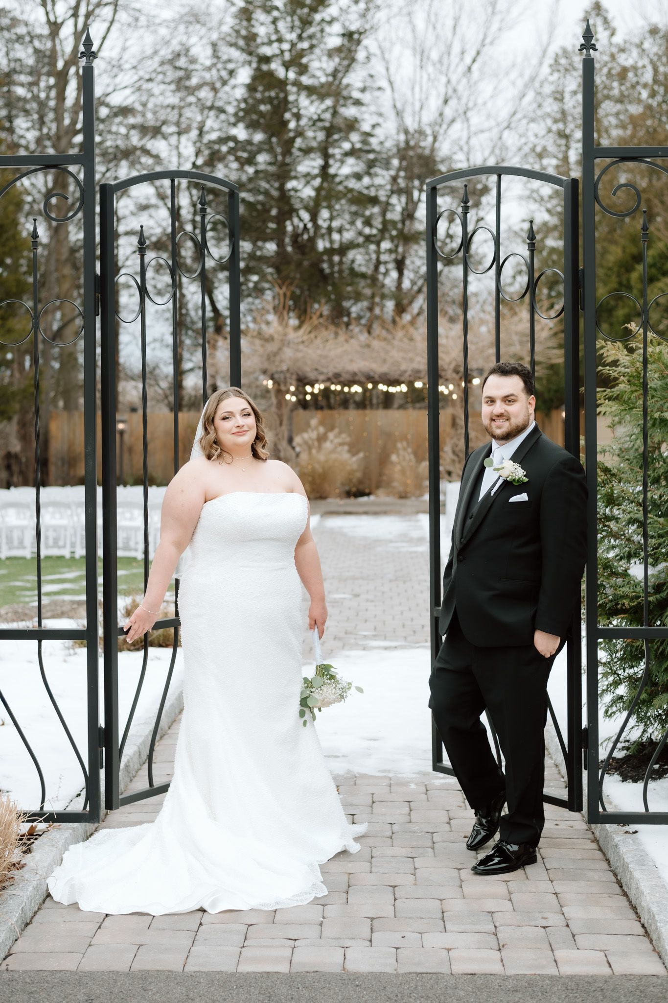 Bride and groom pose at ornate iron gates in a snowy winter garden setting.