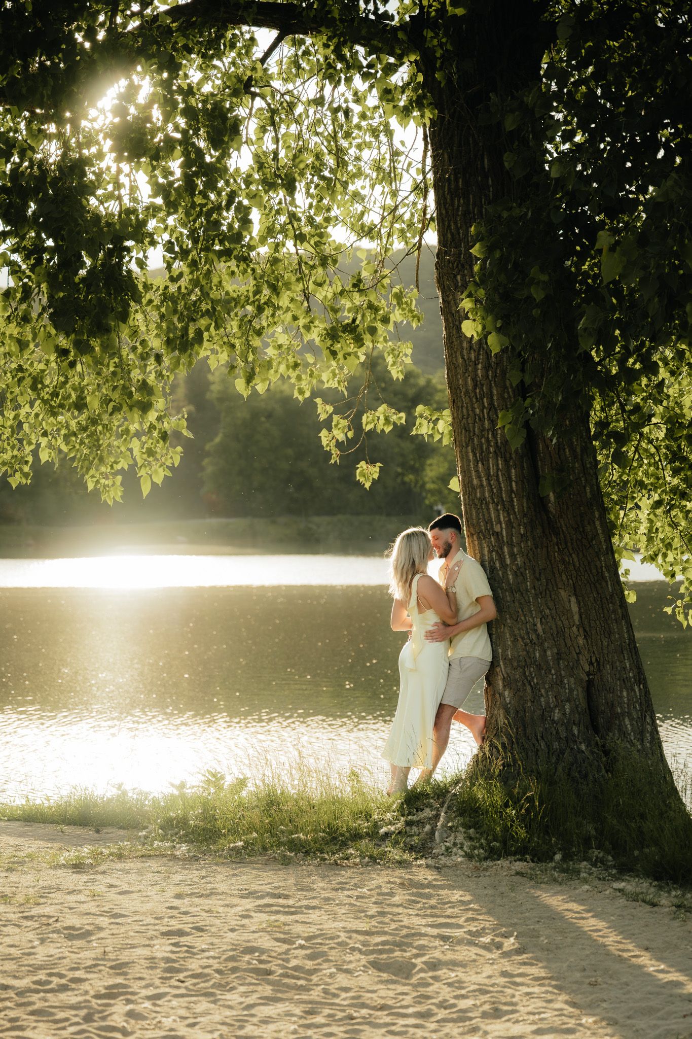 Romantic couple kissing under a large tree by a sunlit lake on a sandy shore in golden hour light.