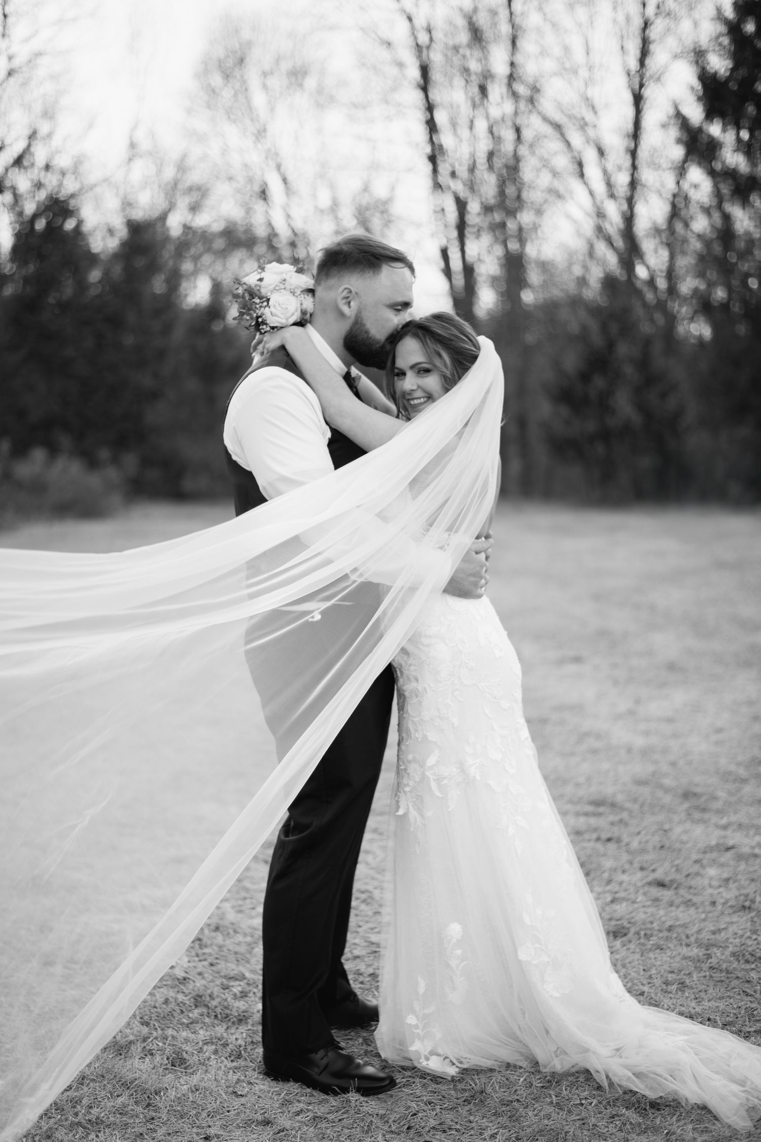 Black and white portrait of groom embracing bride with flowing veil in outdoor setting.