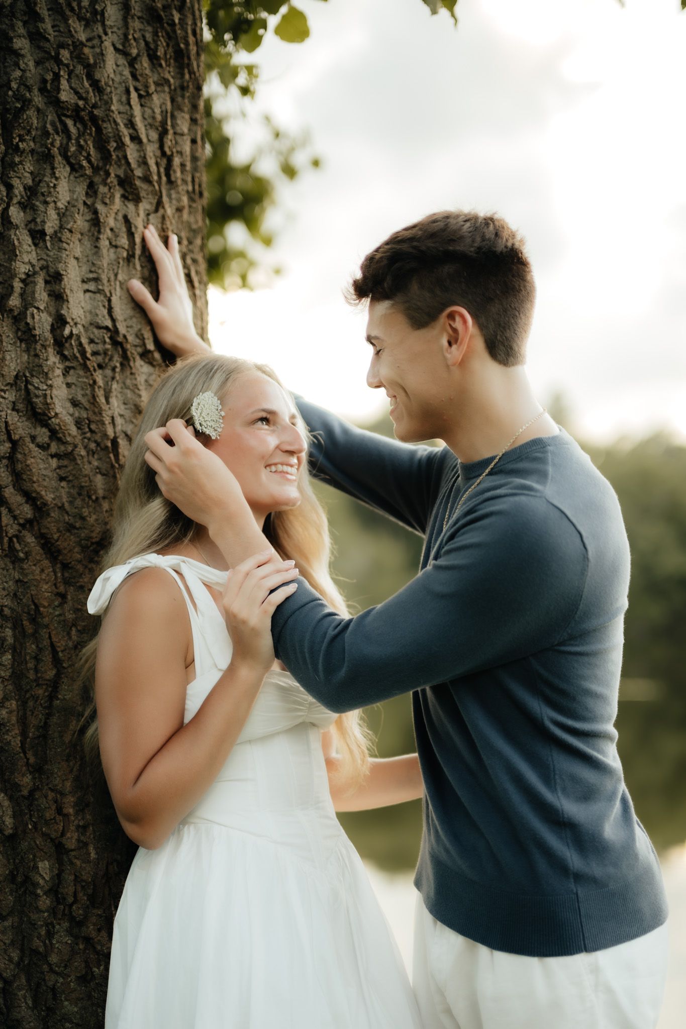 A couple shares a romantic moment by a tree, with the man gently tucking hair behind the smiling woman's ear outdoors.