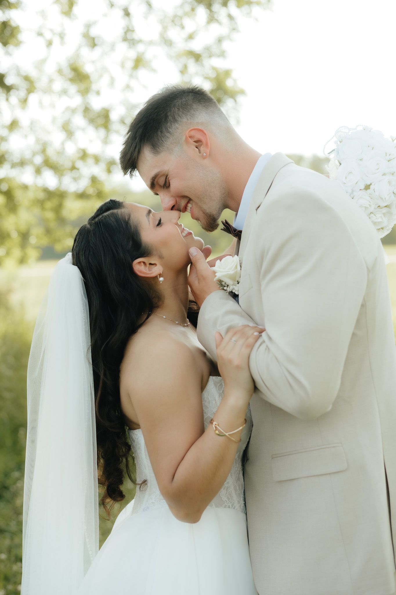 Bride and groom sharing a romantic kiss outdoors, wearing white wedding dress and beige suit, surrounded by lush greenery.