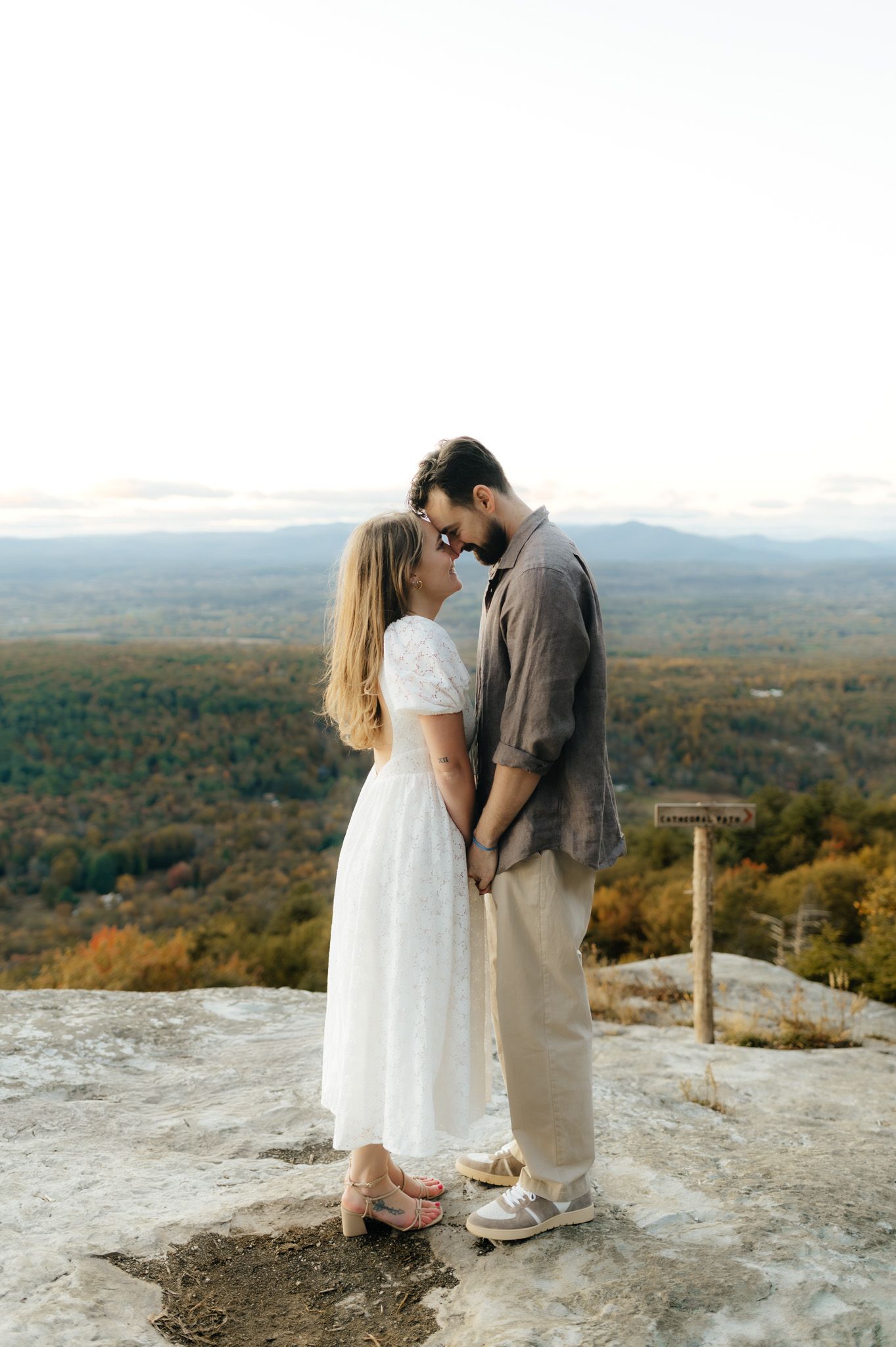 Couple holding hands and touching foreheads on a rocky overlook with stunning autumn mountain views behind them.
