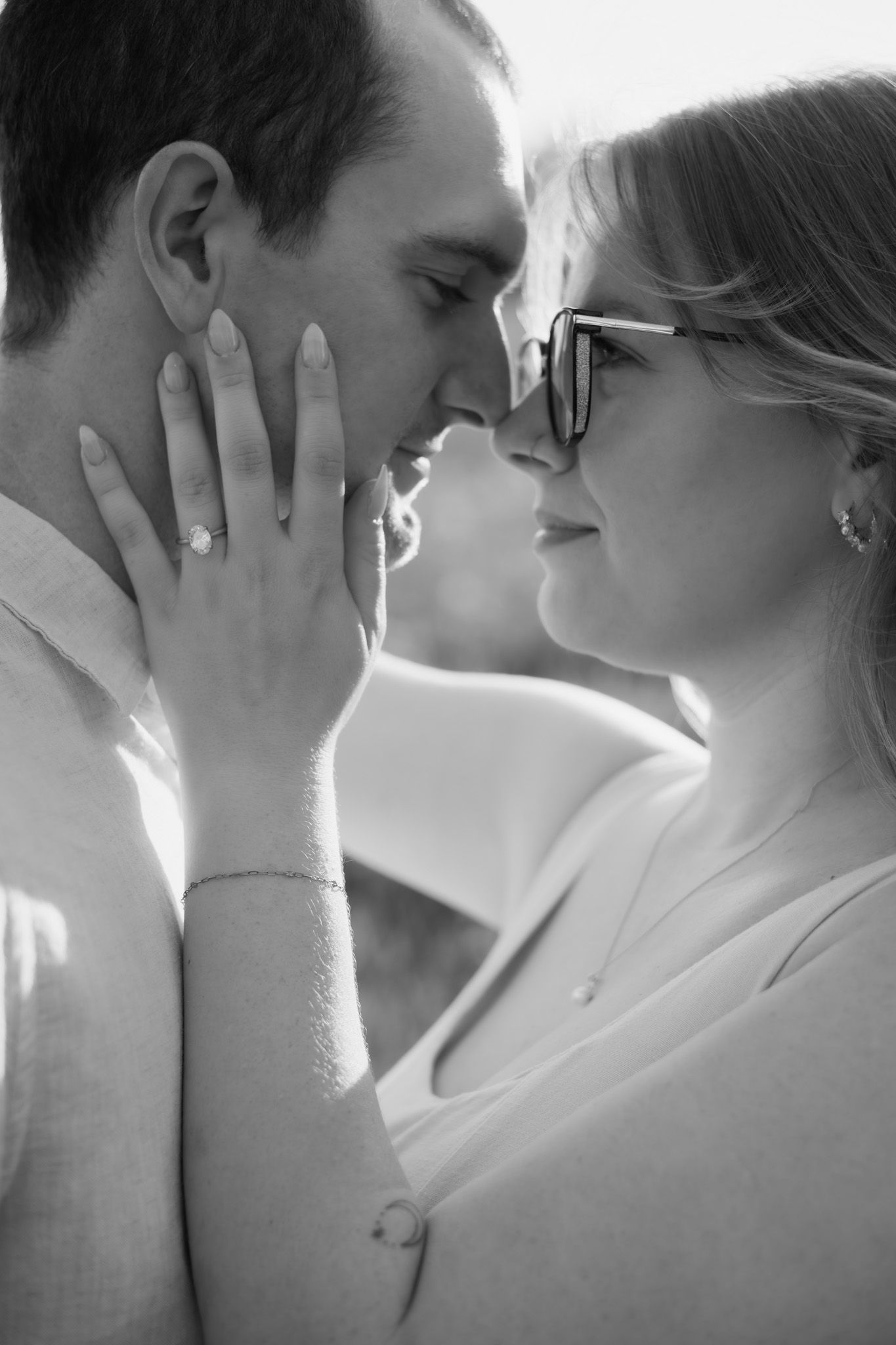 Black and white photo of a couple sharing a romantic moment, woman wearing glasses and engagement ring touching mans face.