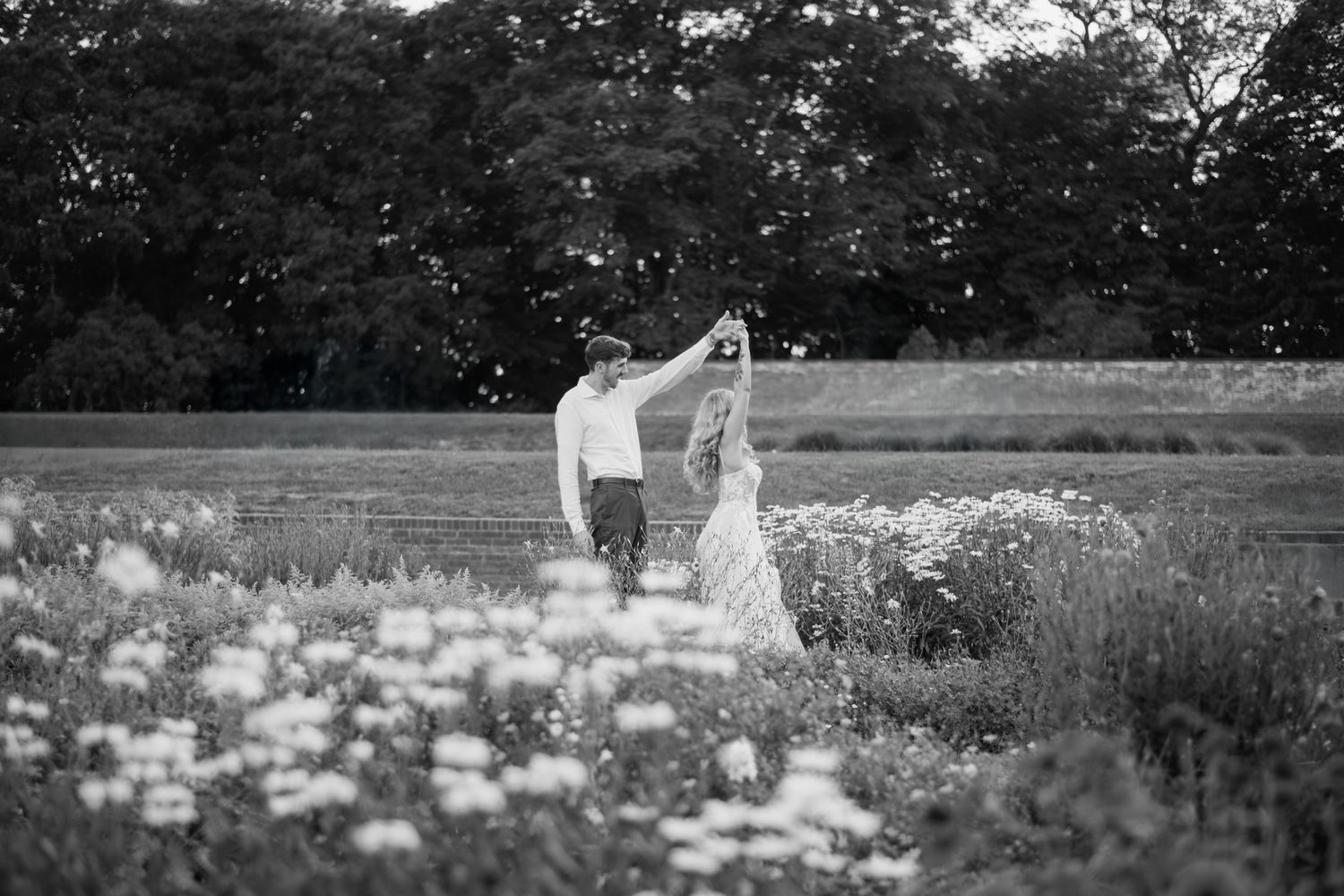 Black and white photo of bride and groom dancing in a wildflower field at dusk.