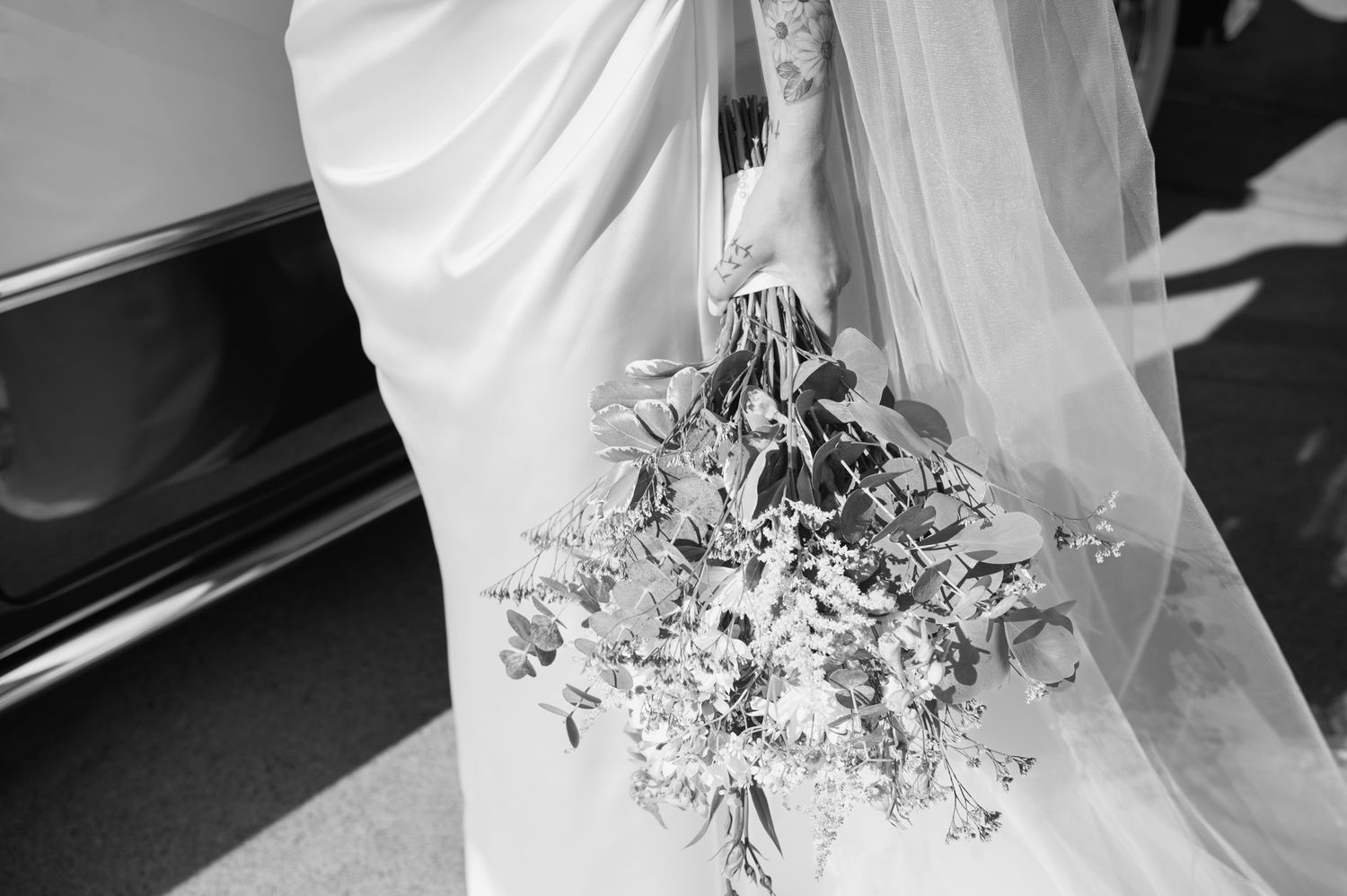 Black and white photo of bride holding a wildflower bouquet beside a vintage car.