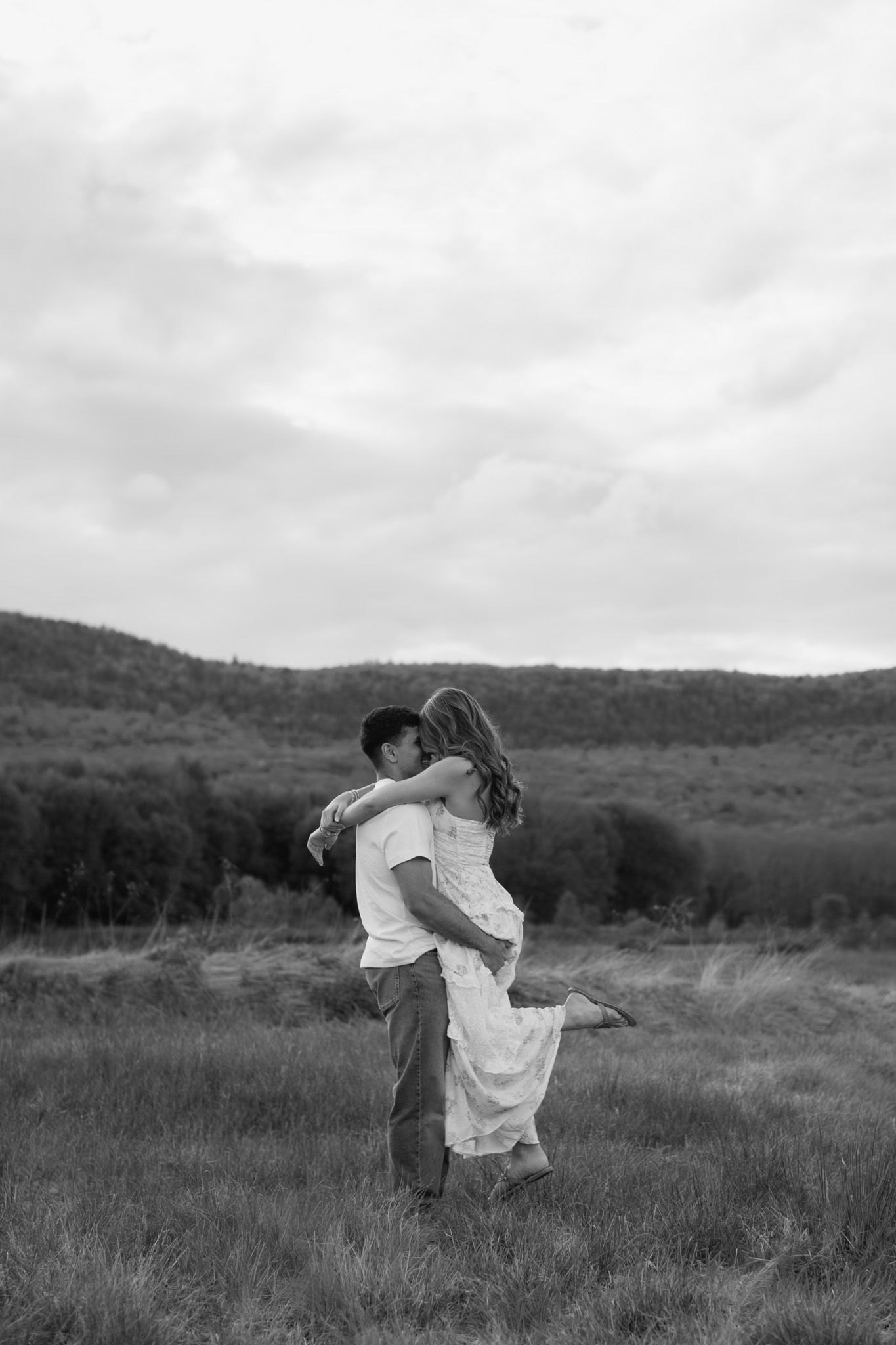 Black and white photo of a man lifting a woman in a field with rolling hills and cloudy sky backdrop.