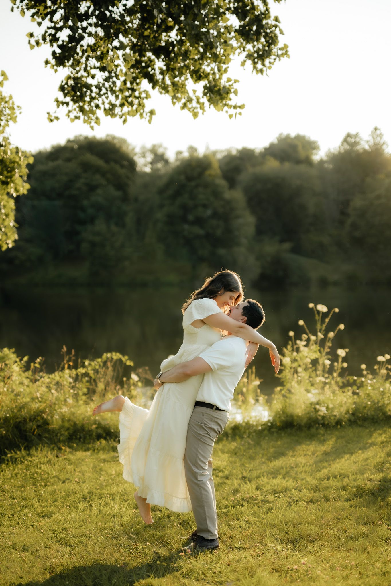Groom lifting bride in white dress by a sunlit lake surrounded by lush green trees on a warm summer day.
