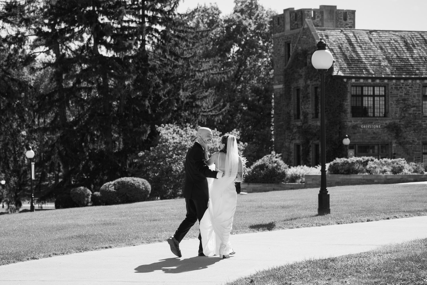 Bride and groom walking hand in hand along pathway near historic stone building in black and white.