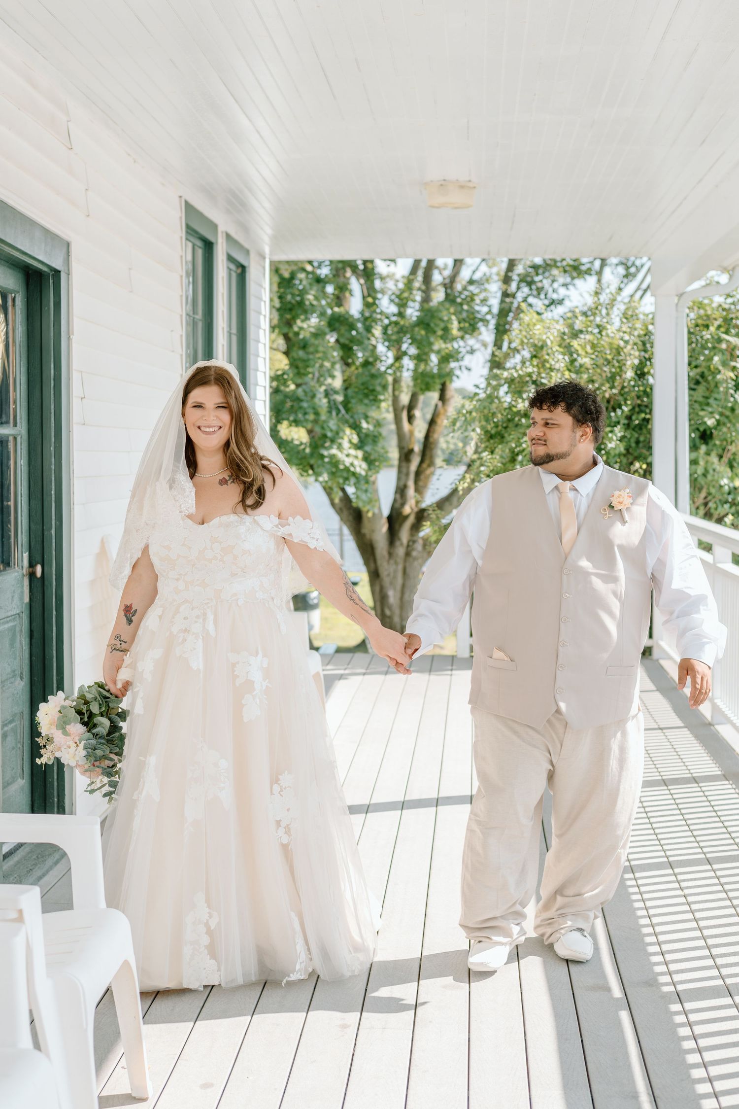 Smiling bride in lace gown and groom in tan suit holding hands on a sunlit white porch.