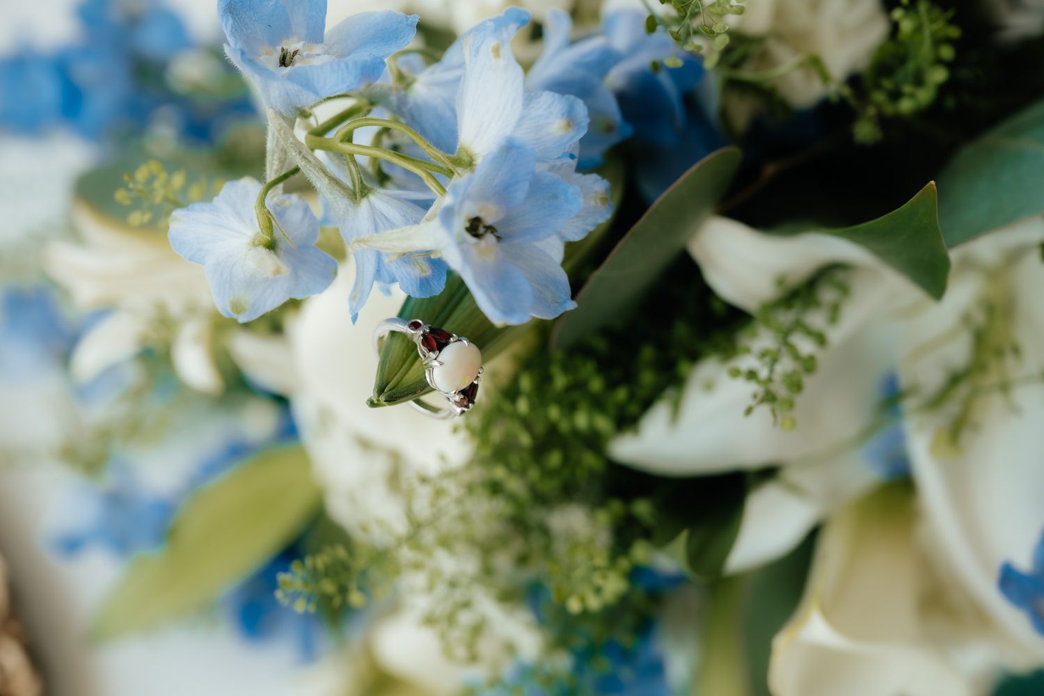 Close-up of a wedding ring nestled among blue delphinium, white orchids, and green flowers in an elegant bridal bouquet.