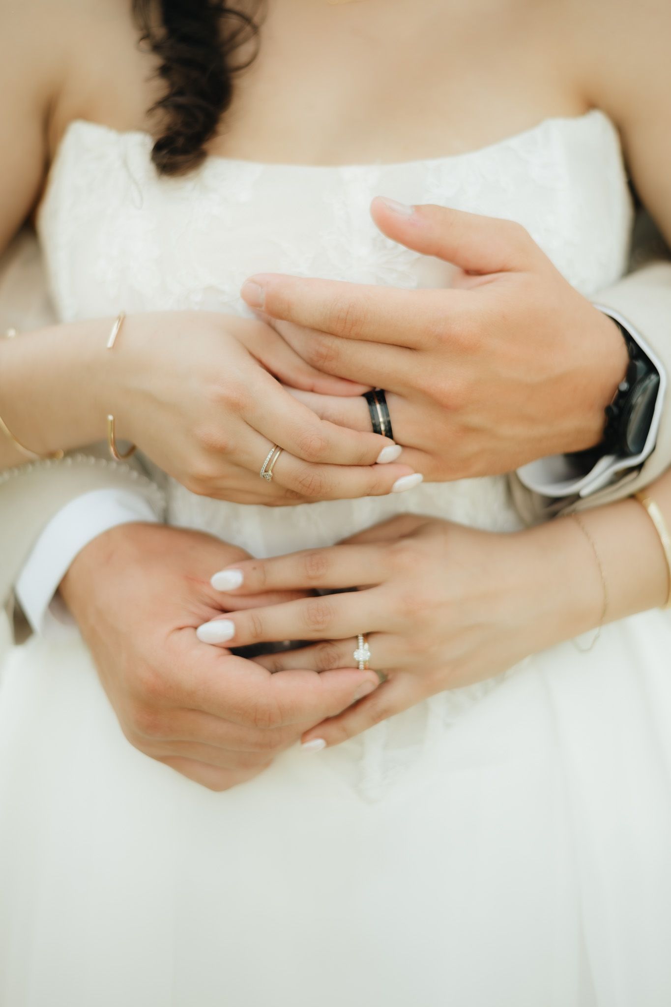 Bride and groom holding hands showing wedding rings, white dress and suit, diamond engagement ring and black band.