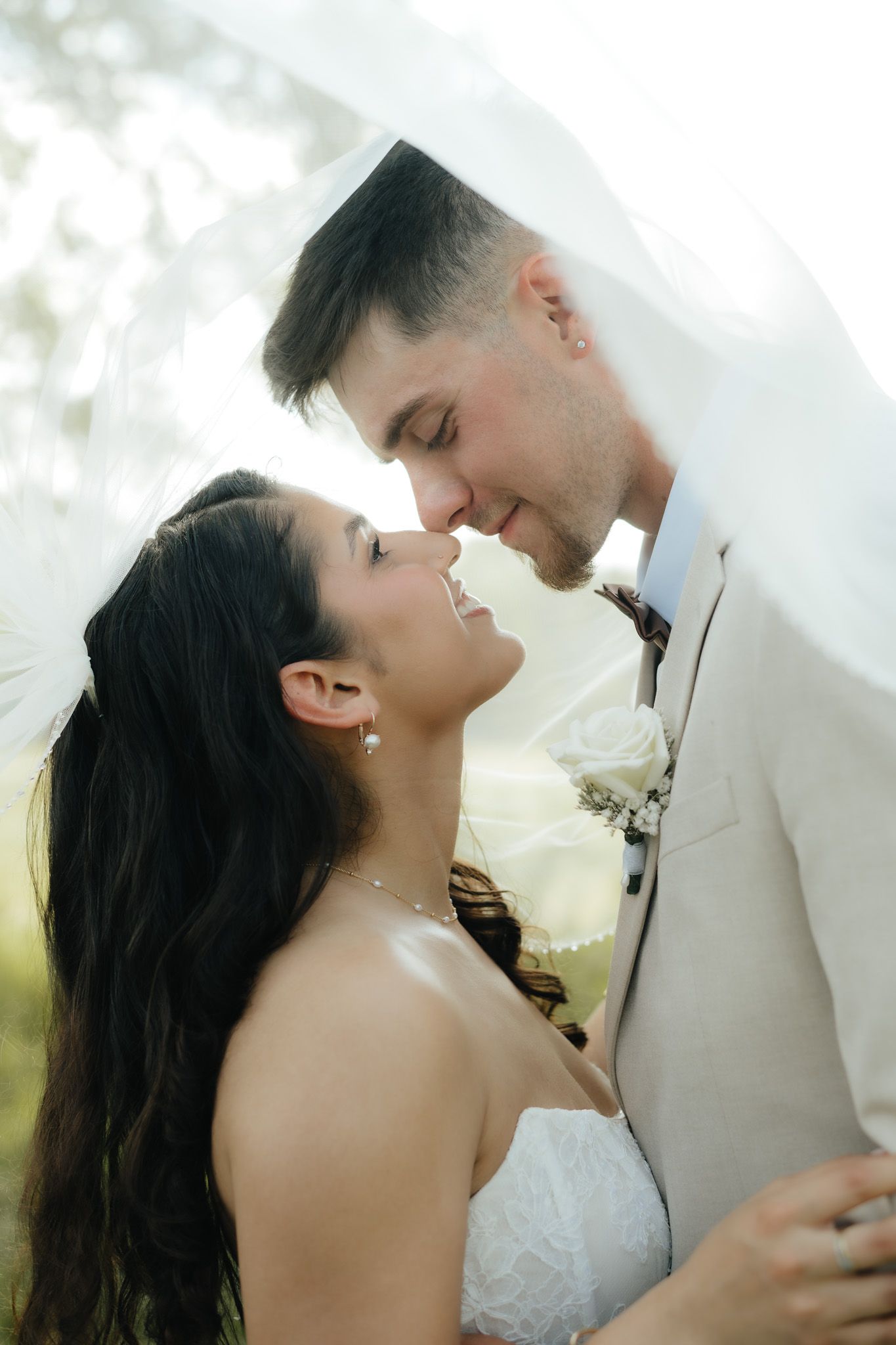 Bride and groom share a romantic moment under a flowing veil, dressed in wedding attire, smiling at each other outdoors.