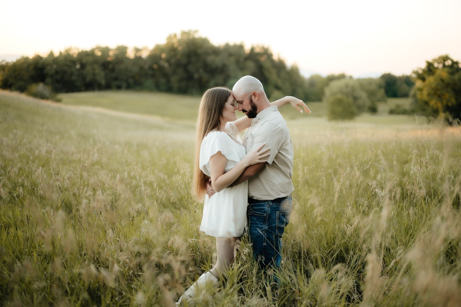 A couple sharing a romantic moment in a sunlit green meadow, the man kissing the woman in a white dress.