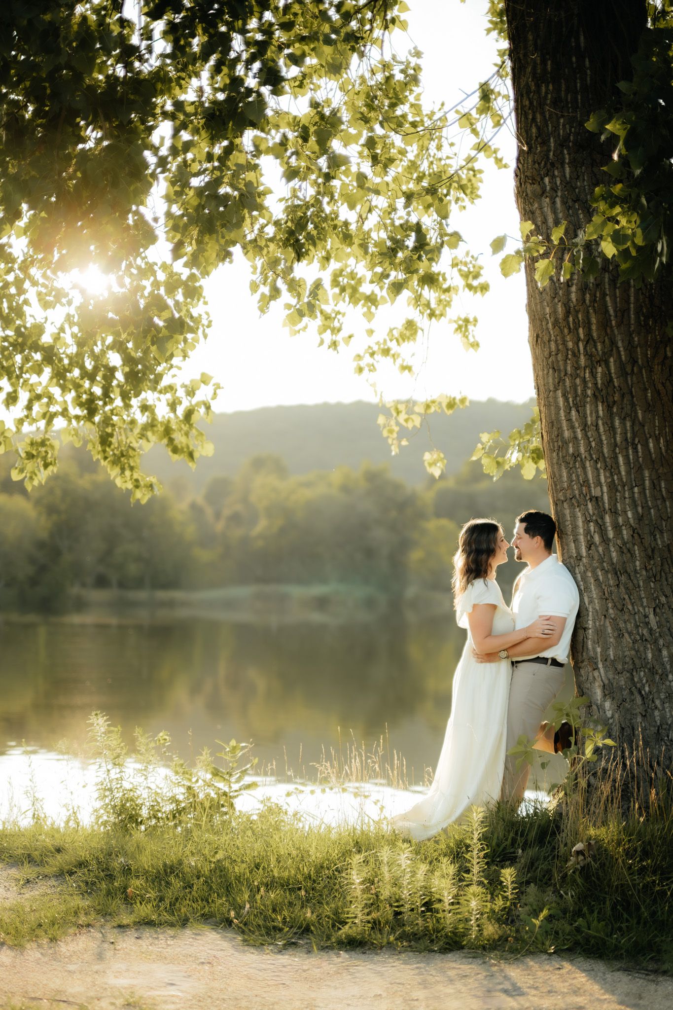 Couple embracing under a large tree by a sunlit lake at golden hour, bride in white gown, lush greenery background.