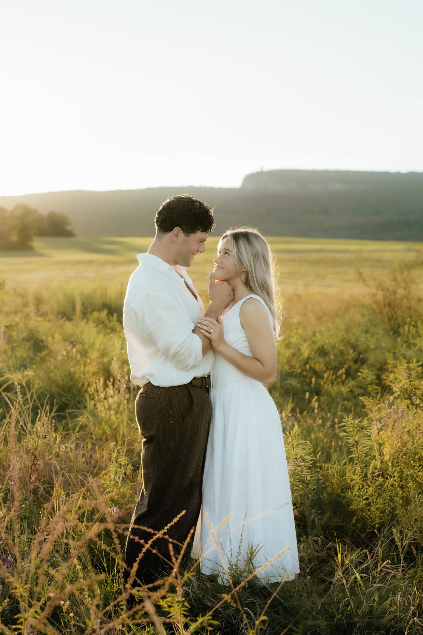 Couple embracing in golden hour sunlight in a lush green field, woman in white dress smiling up at man in white shirt.