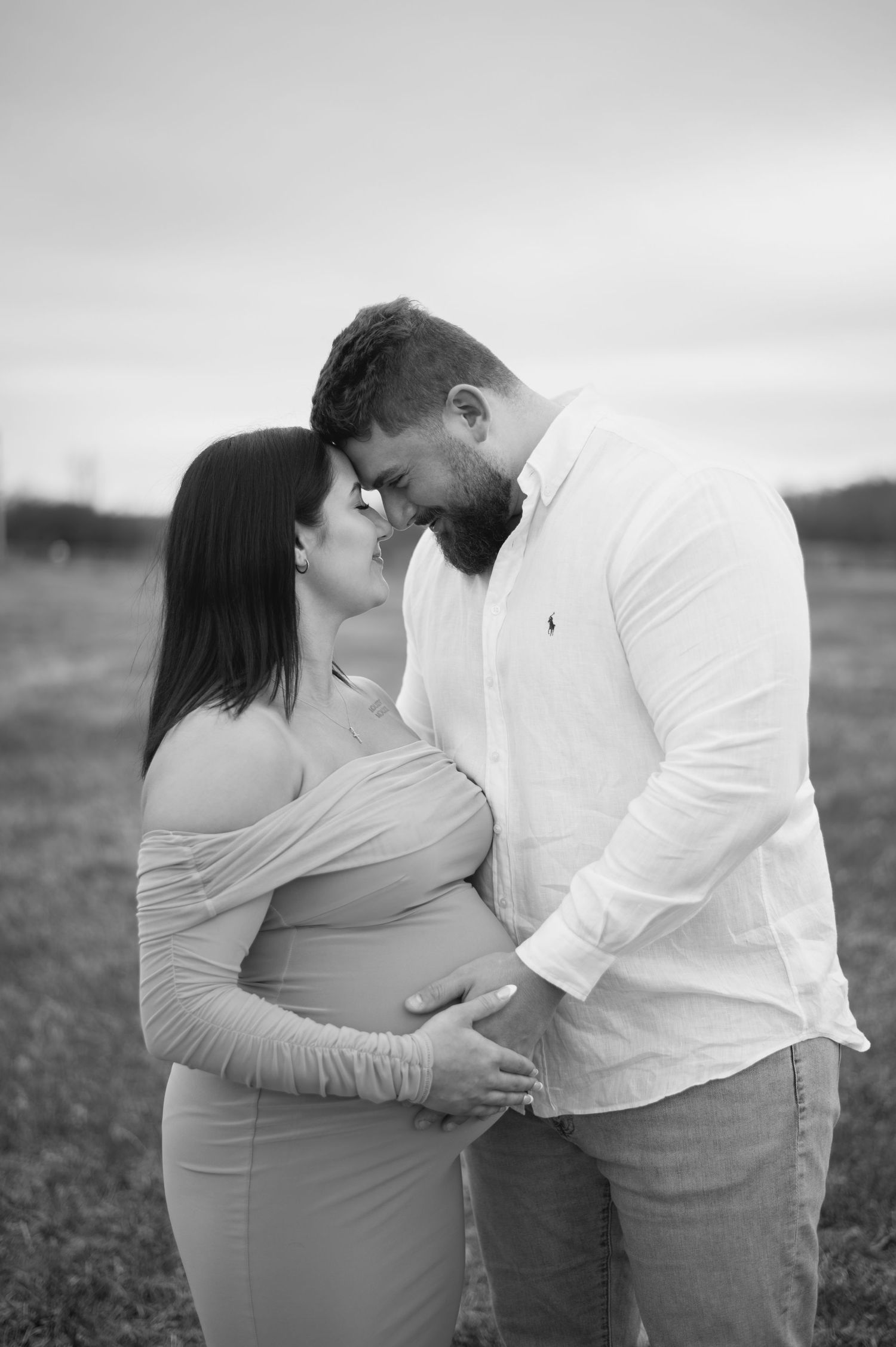 Black and white maternity photo of a couple touching foreheads in a field, cradling the pregnant belly.