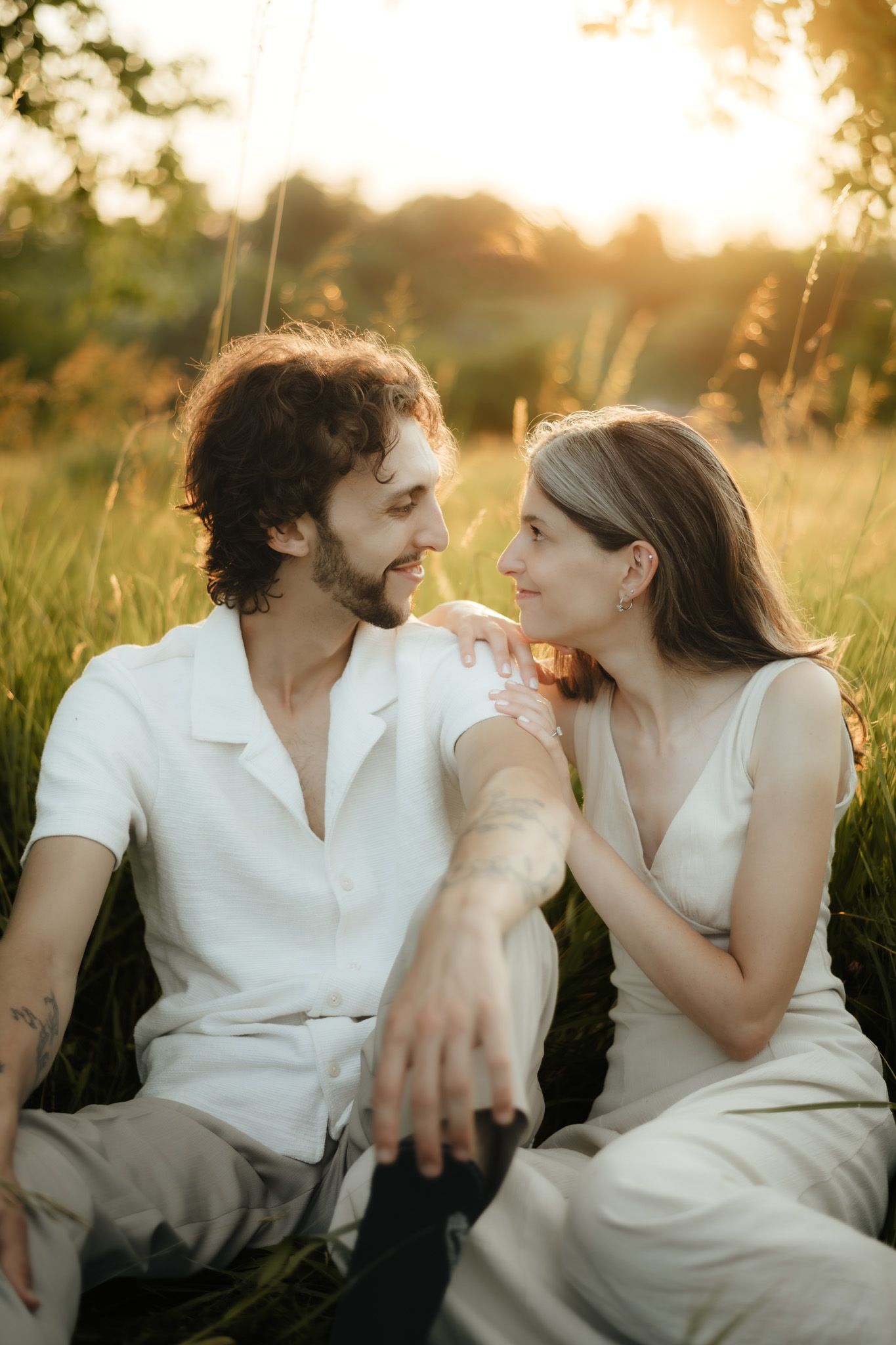 Couple sitting in golden sunset field, wearing white outfits, gazing at each other romantically.