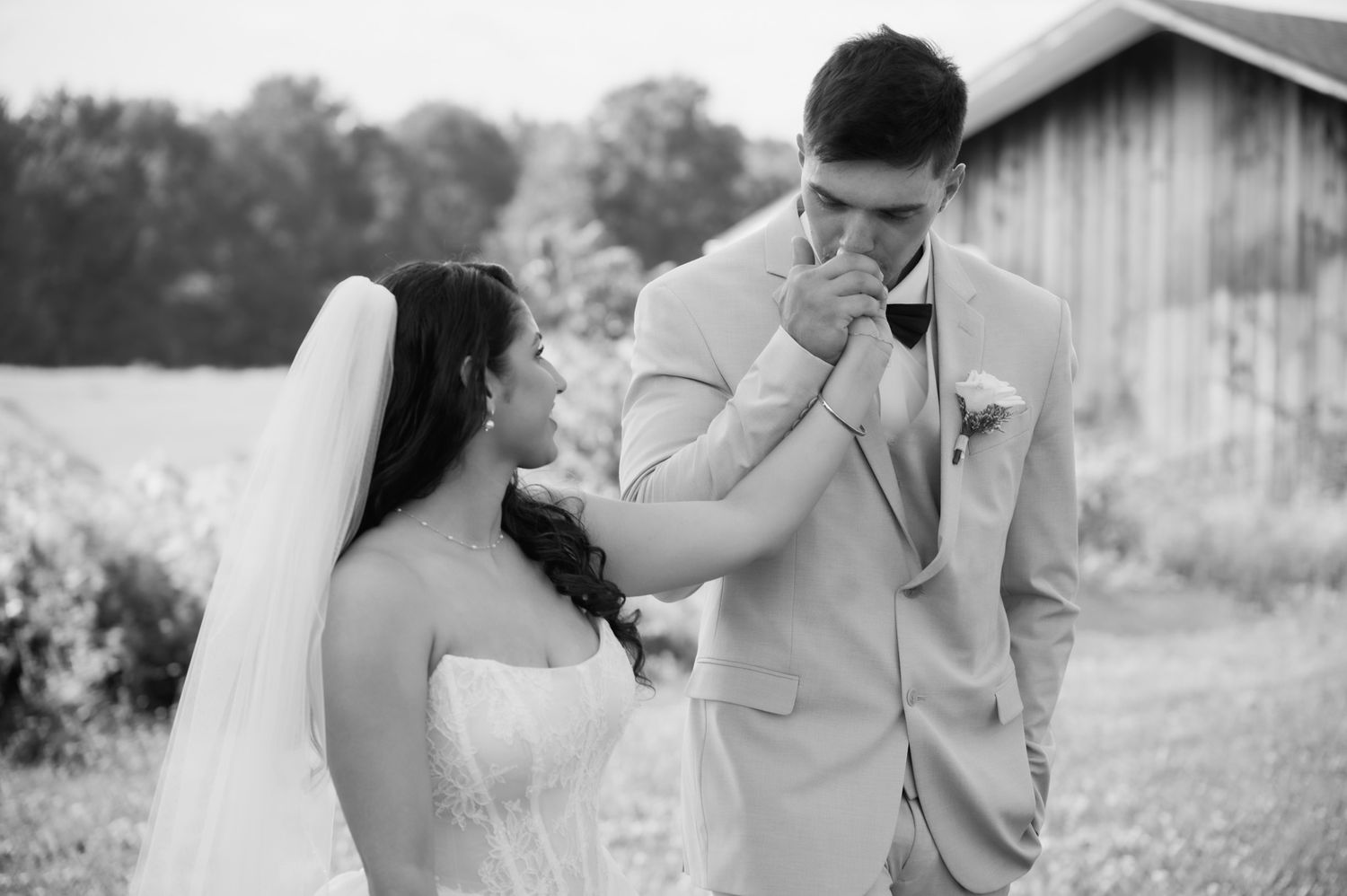Groom kissing bride's hand in a romantic black and white outdoor wedding photo near a rustic barn.