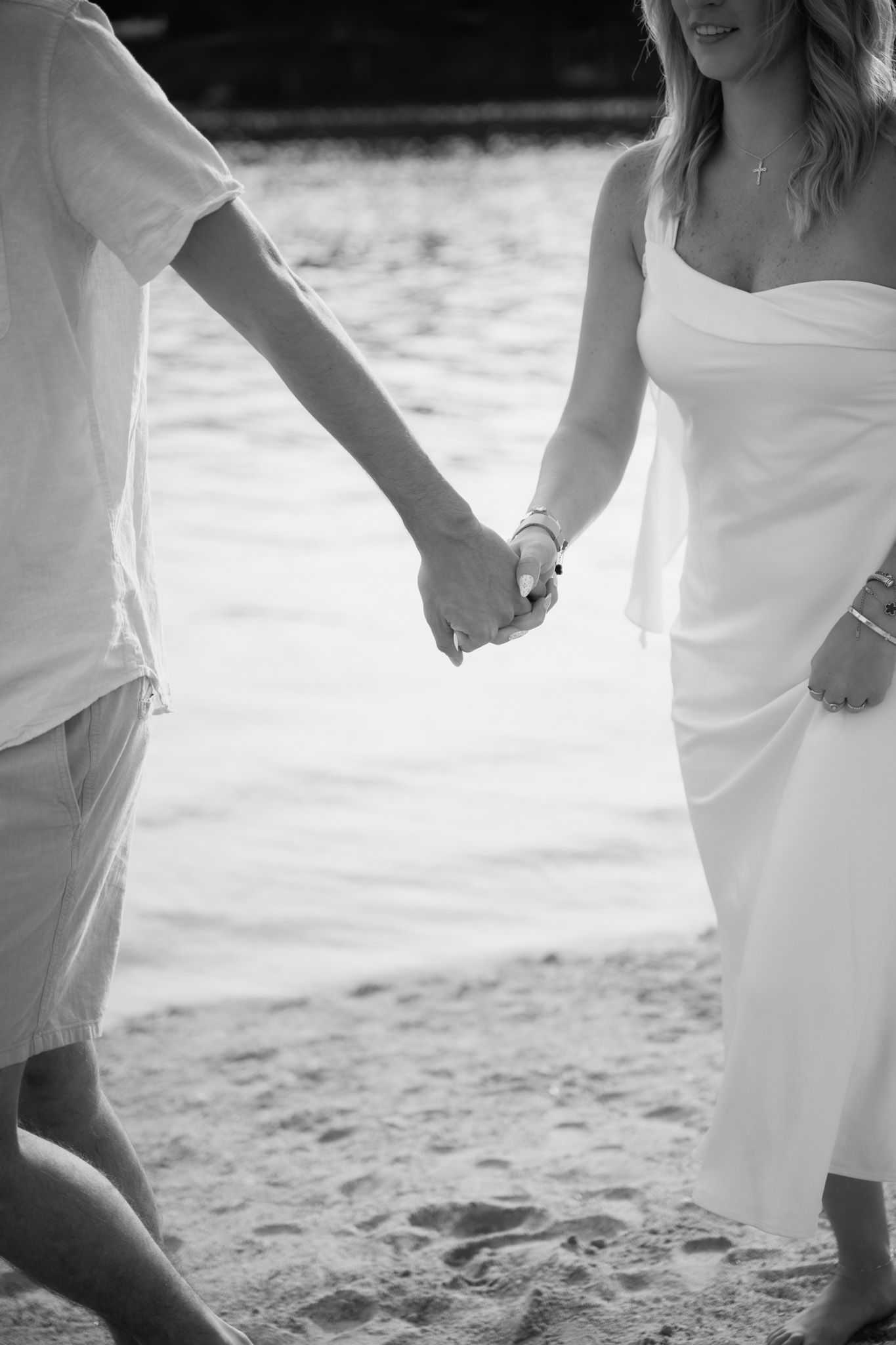 Black and white photo of a couple holding hands on a sandy beach, woman in white dress, man in white shirt.