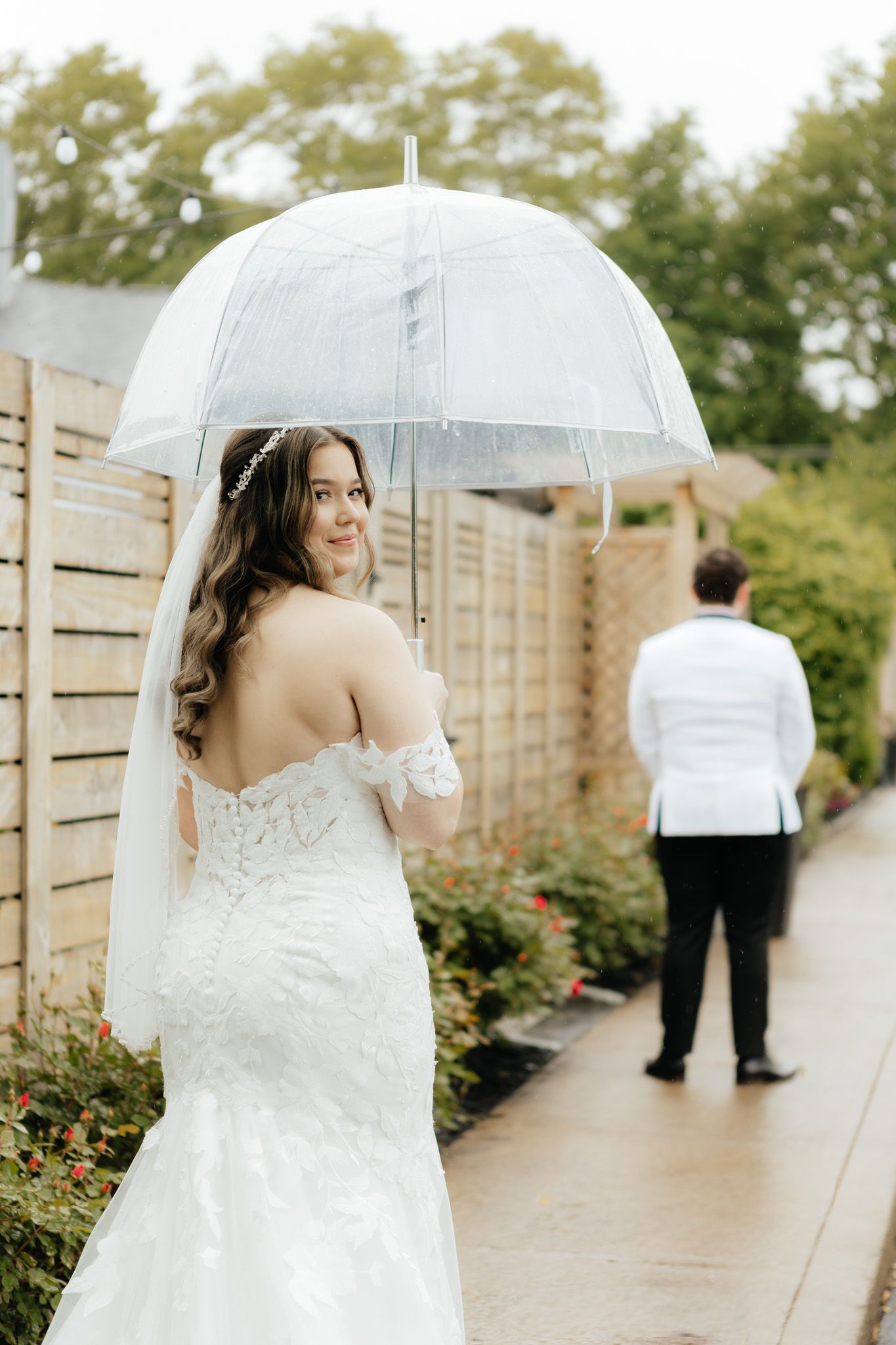 Bride in lace off-shoulder gown holding a clear umbrella, smiling over her shoulder on a rainy wedding day.