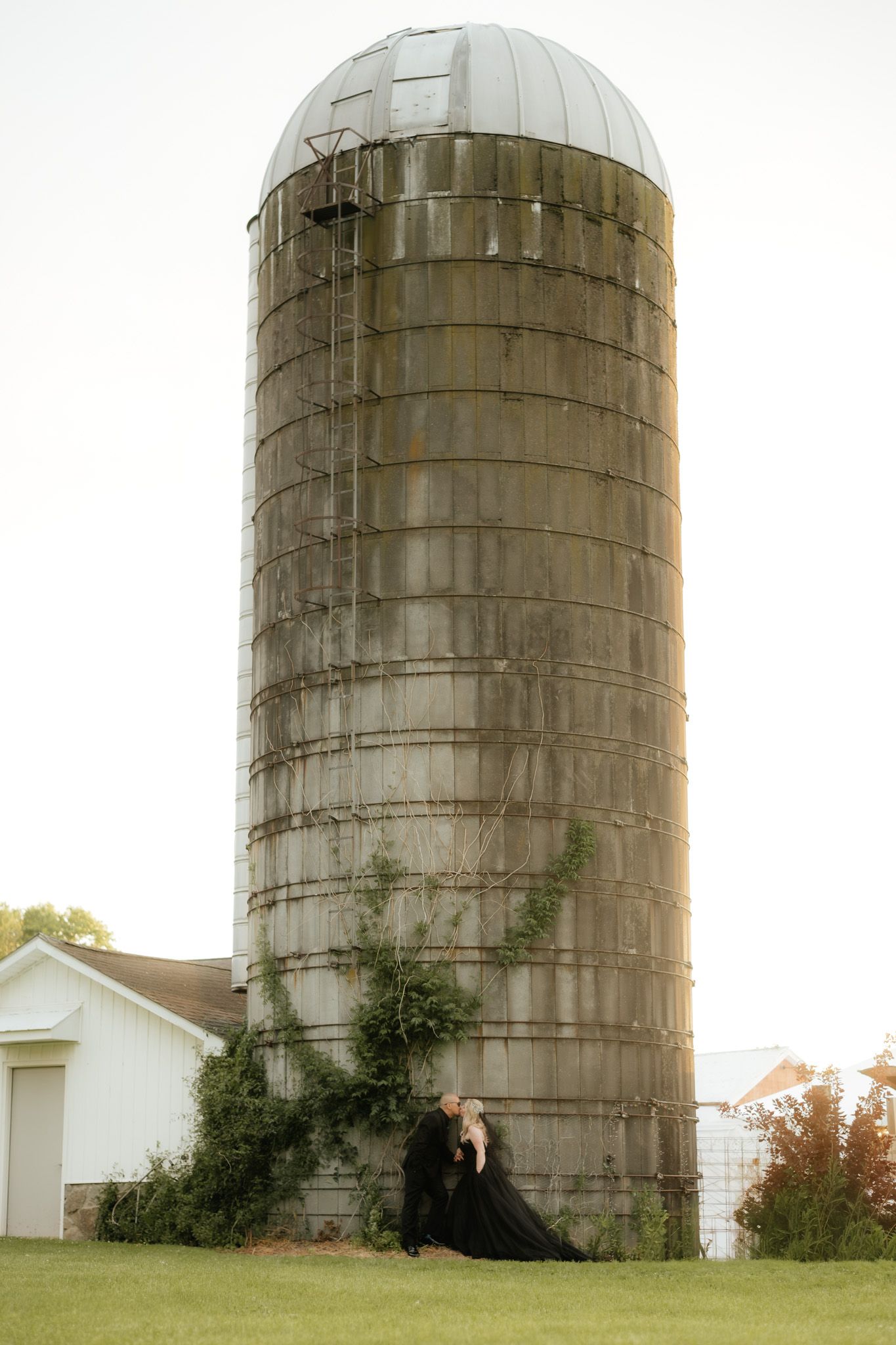 Couple in black attire posing romantically beside a rustic farm silo with ivy, surrounded by lush greenery.