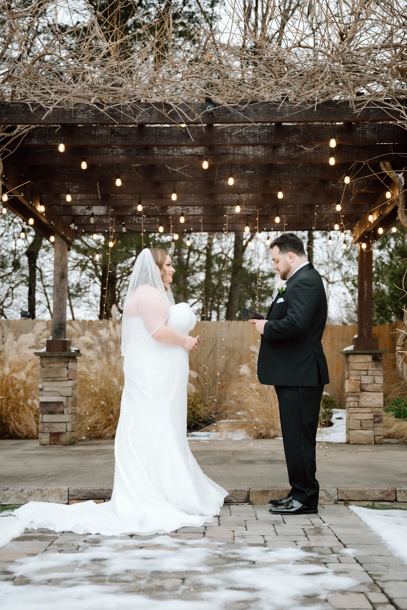 Bride and groom exchange vows under a pergola with string lights in the snow.