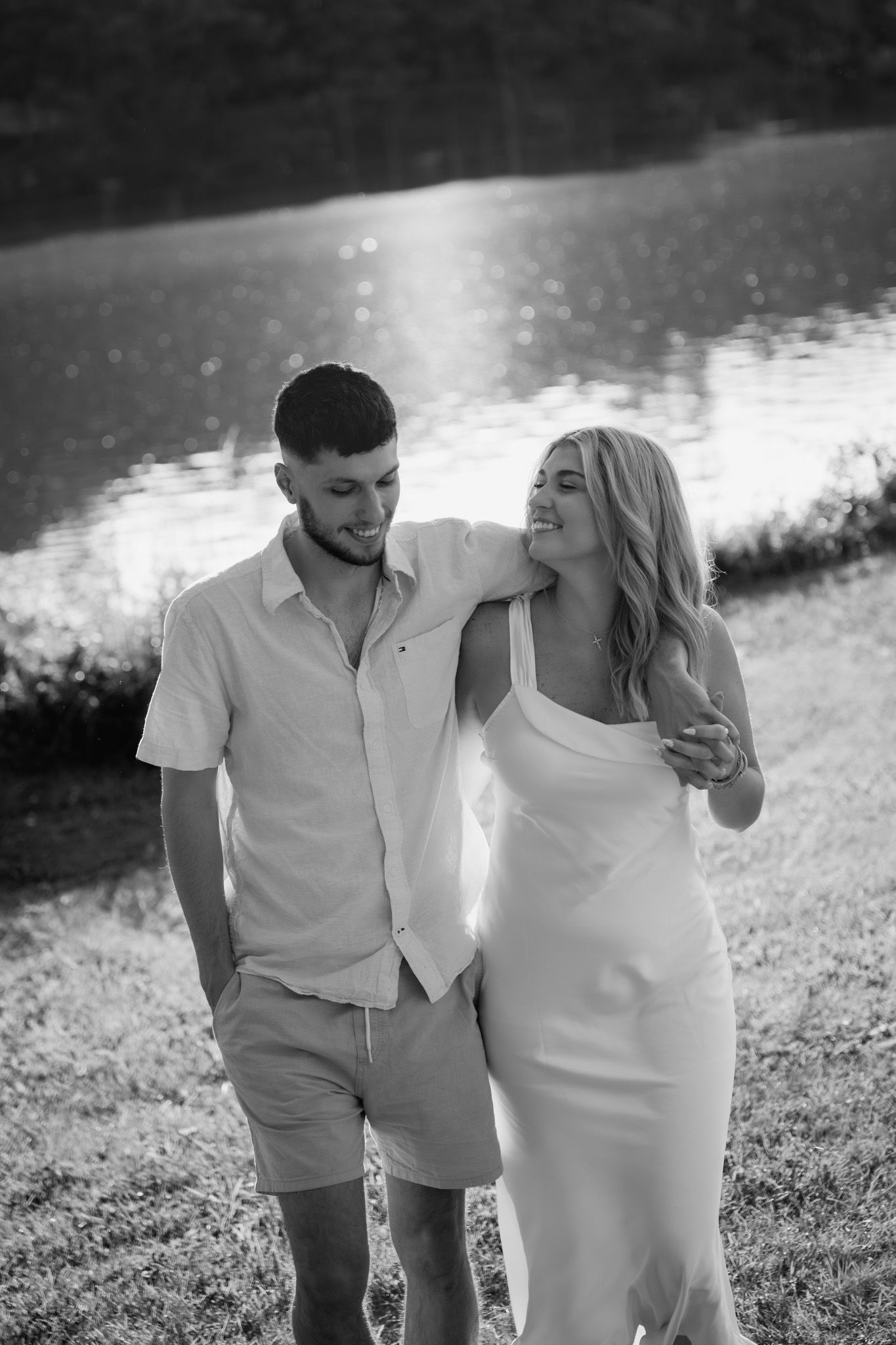 Black and white photo of a couple walking by a lake, woman in white dress with mans arm around her shoulders.