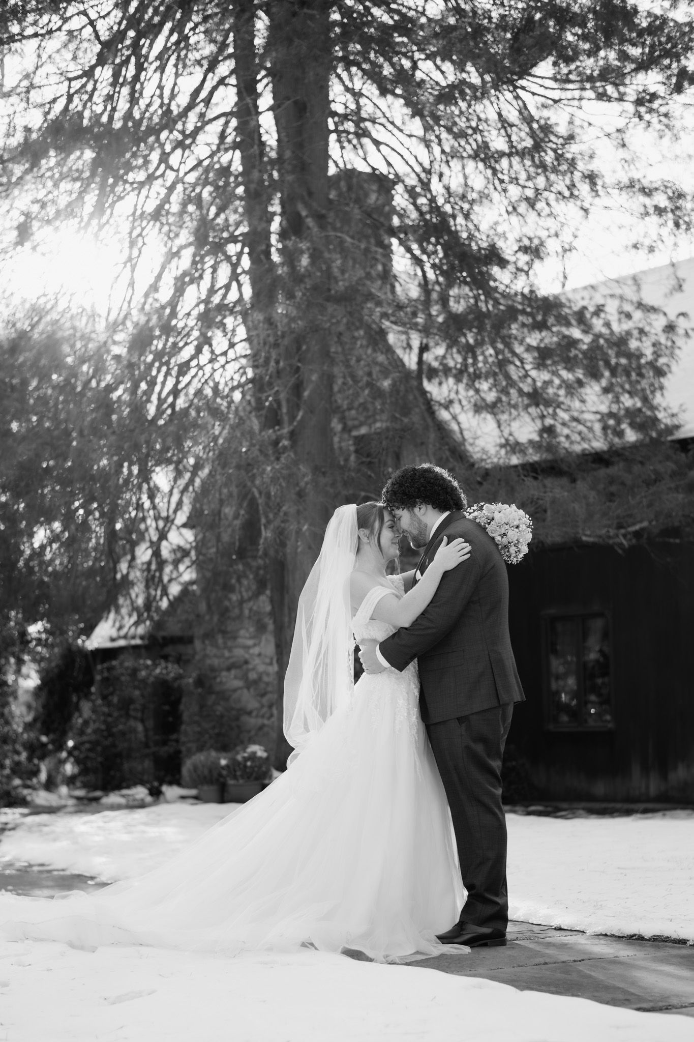 Bride and groom embracing in snowy winter landscape beside historic stone building in black and white.