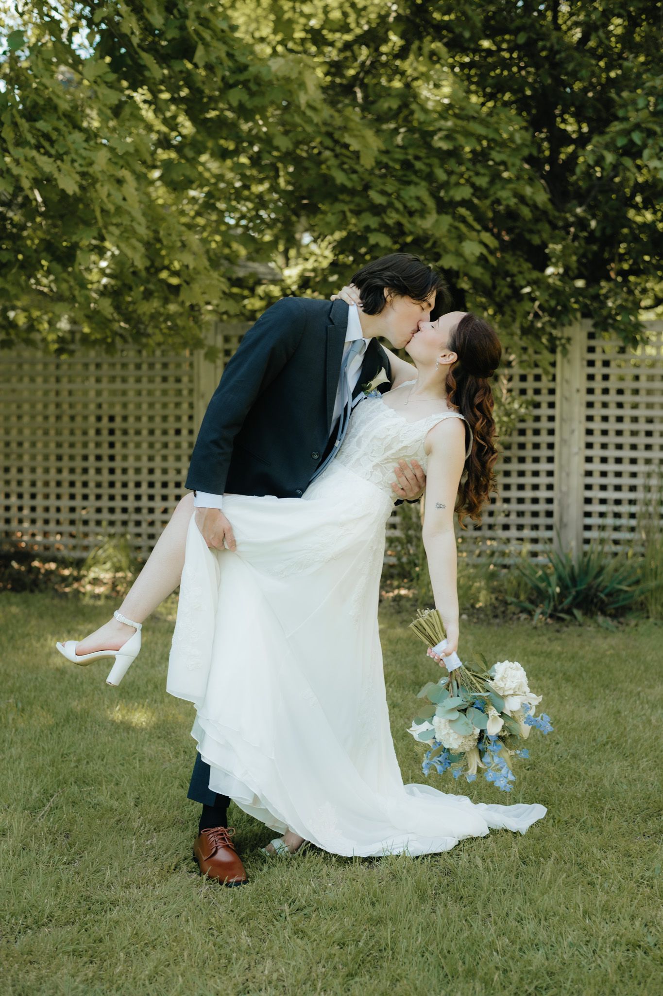 Groom dips and kisses bride in white gown holding blue floral bouquet outdoors on green lawn.