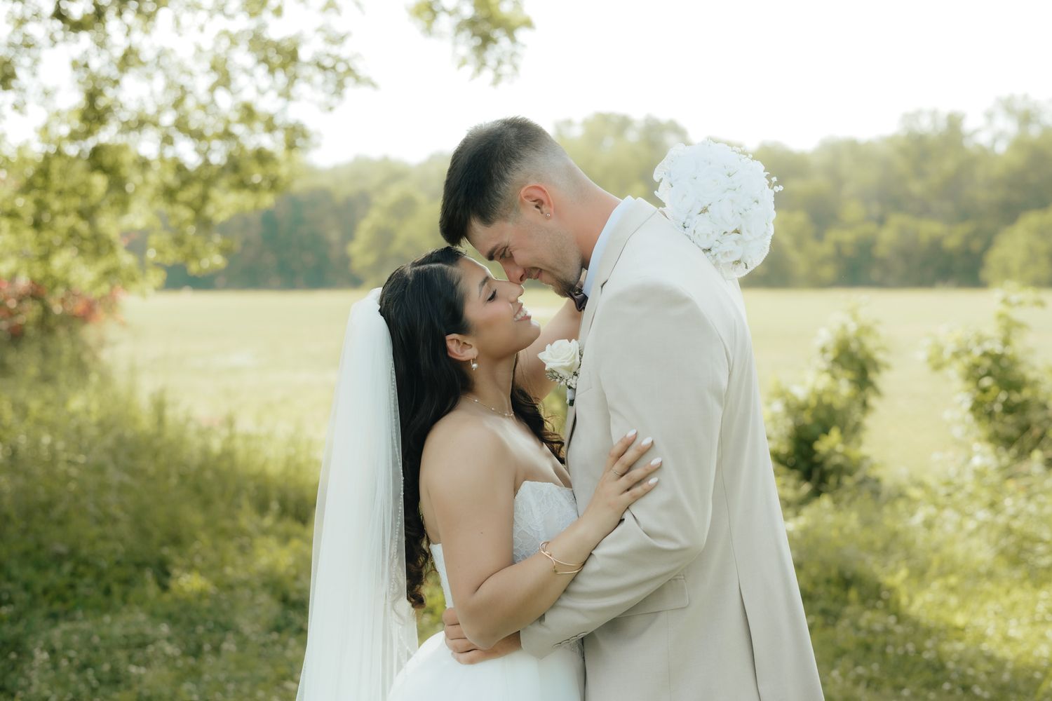 Bride and groom sharing a romantic moment outdoors, embracing in soft sunlight with lush green trees in the background.