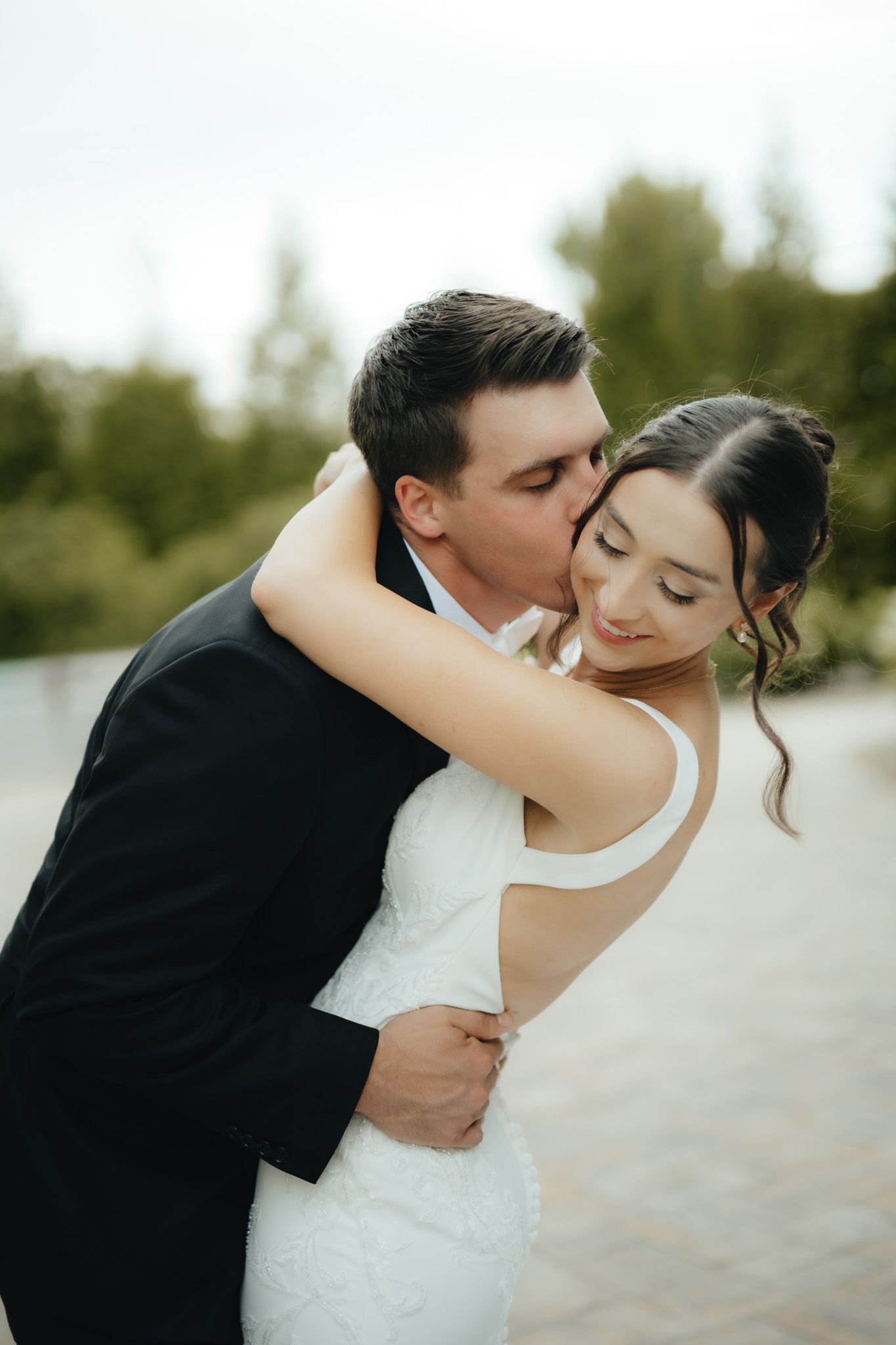 Groom in black suit kisses bride on cheek while embracing her in white wedding dress outdoors.