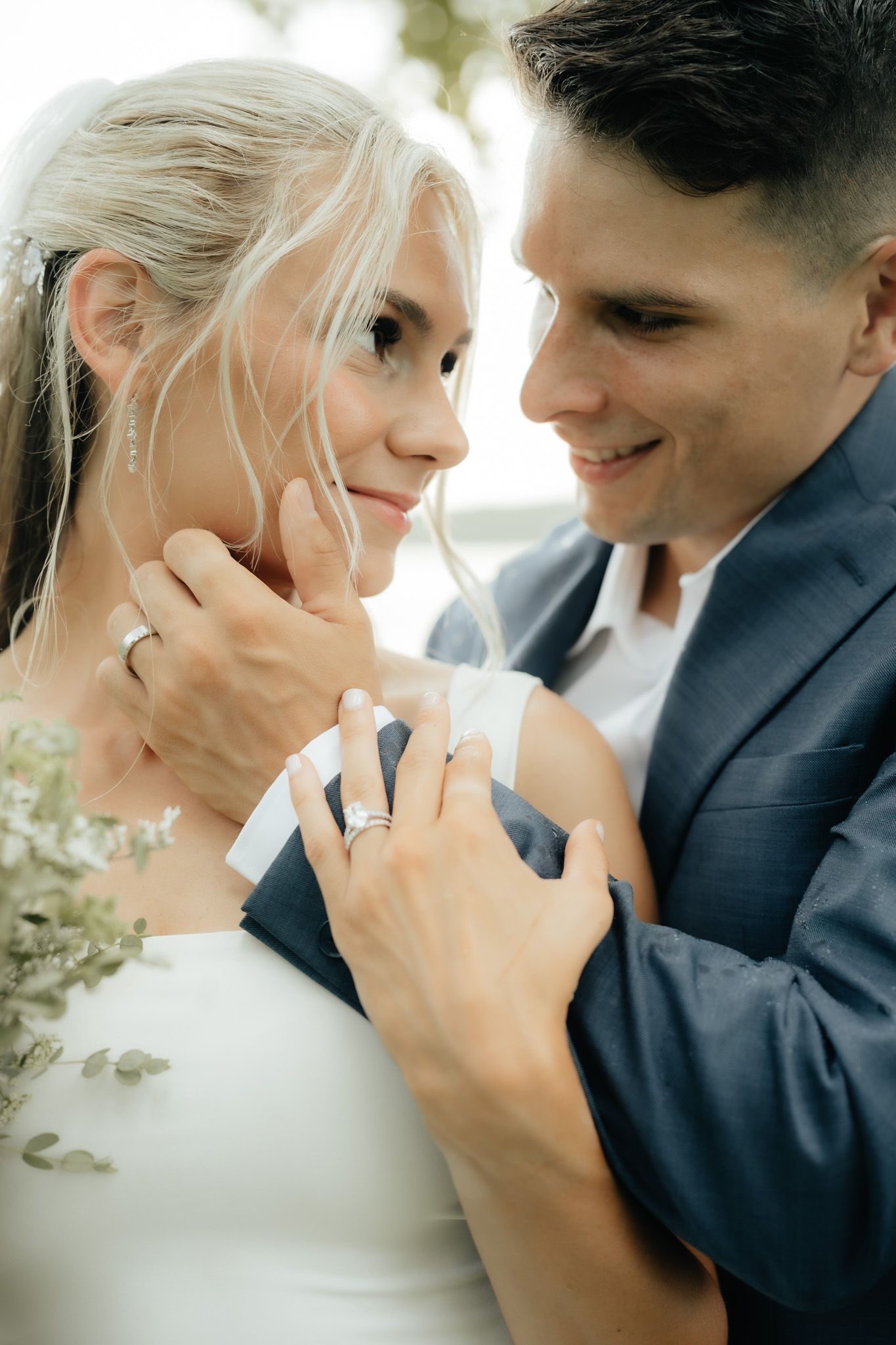 A smiling bride and groom in wedding attire share a tender moment, showcasing their wedding rings.