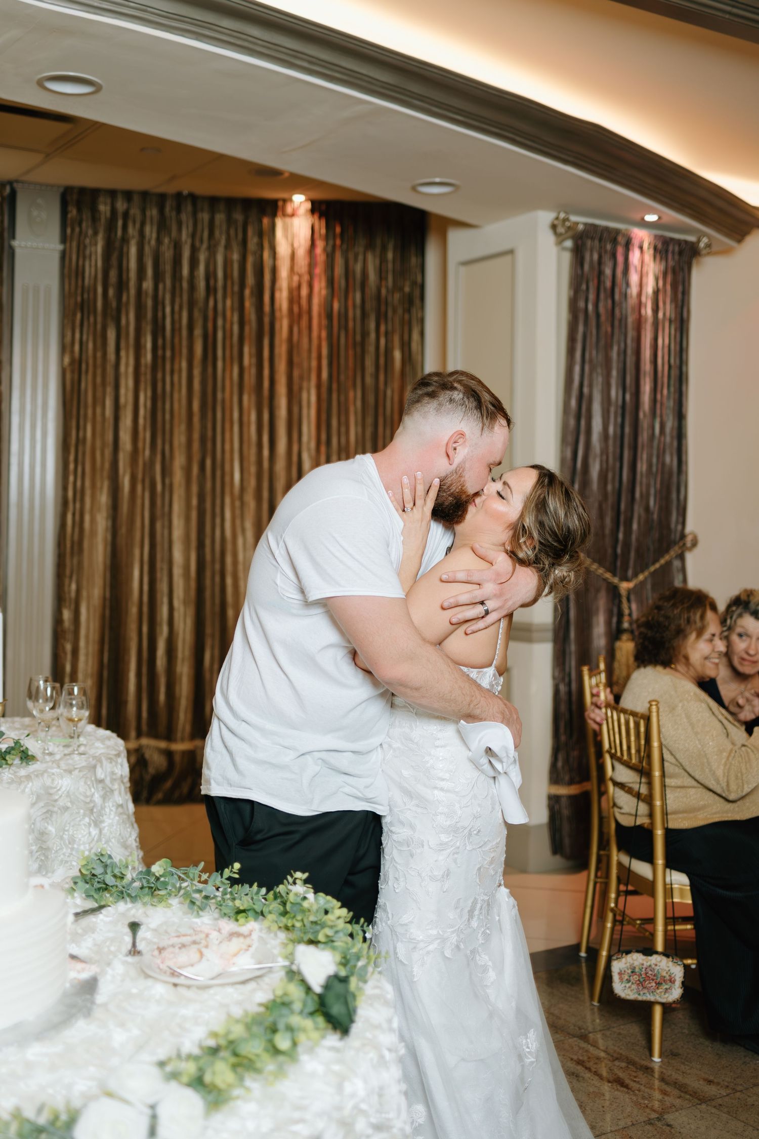 Groom kissing bride near wedding cake during indoor reception celebration with guests nearby.