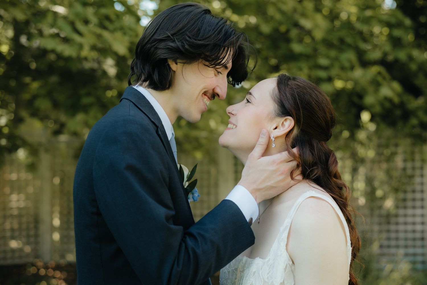 A couple in wedding attire share a tender moment, smiling at each other outdoors with lush green trees behind them.