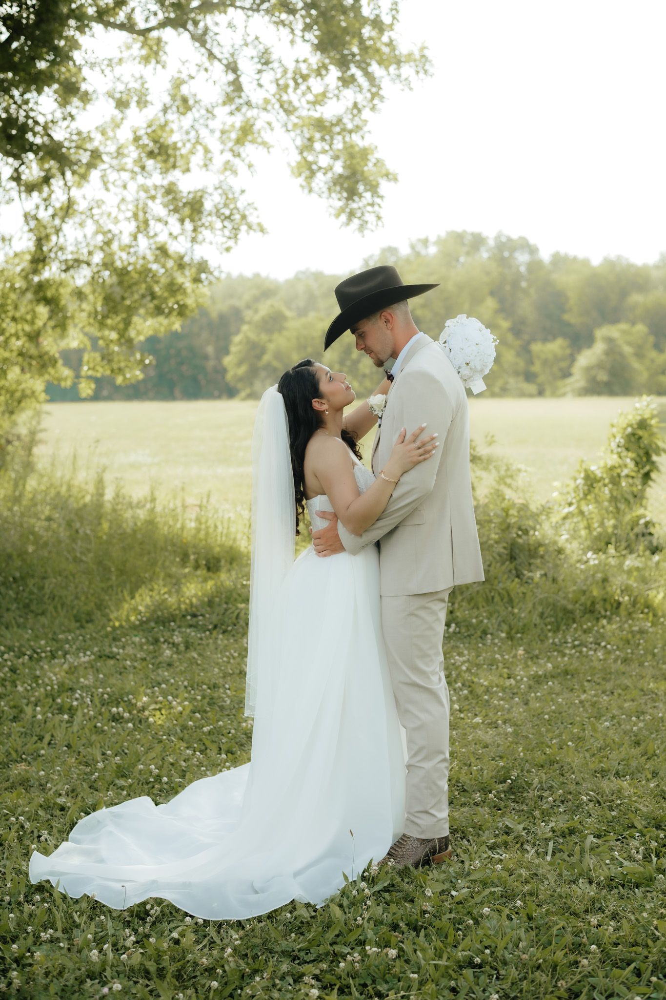 Bride in white gown and groom in tan suit with cowboy hat share romantic moment in lush green outdoor setting.