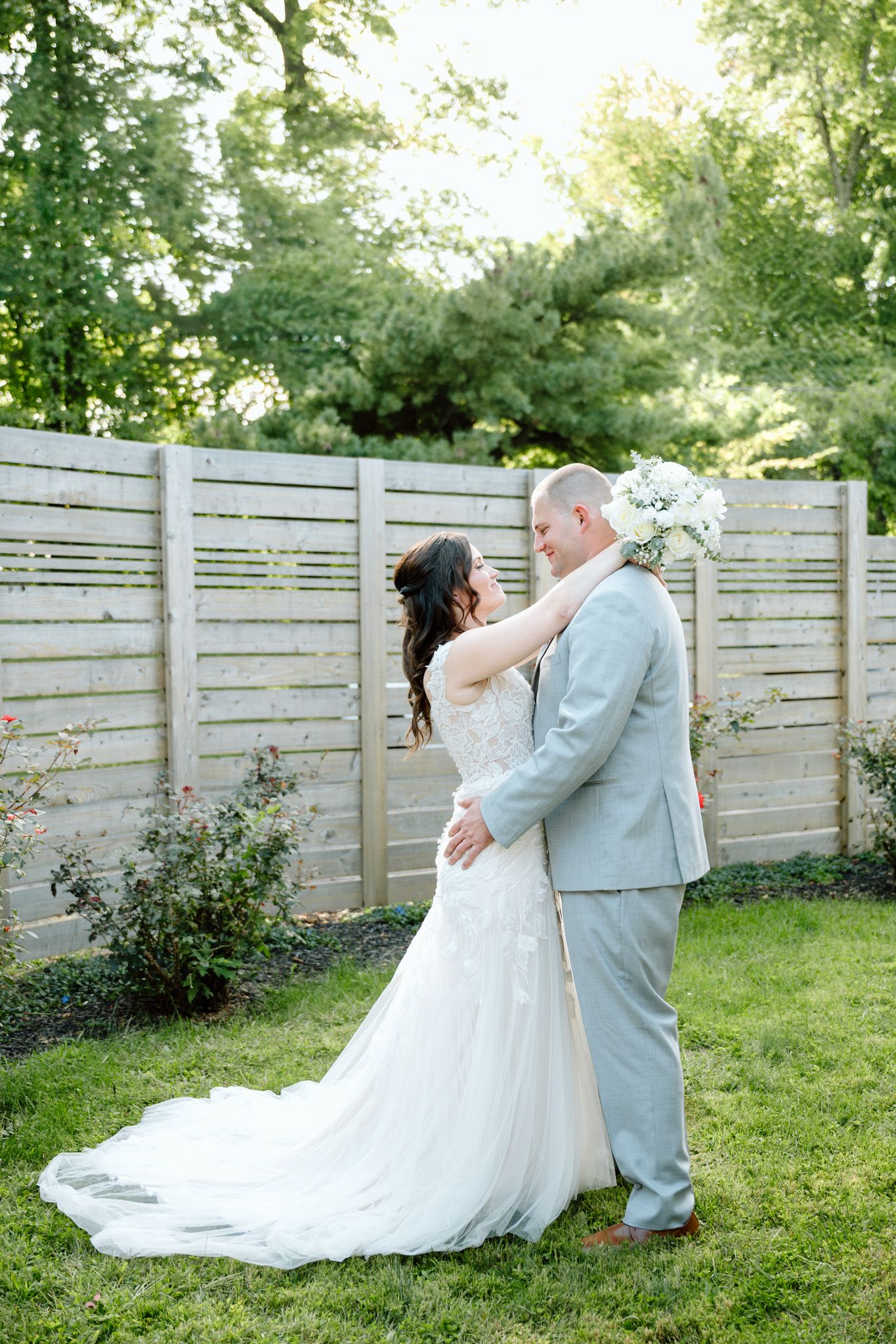 Bride and groom embrace in a lush garden setting, with the bride in a lace gown and long train.