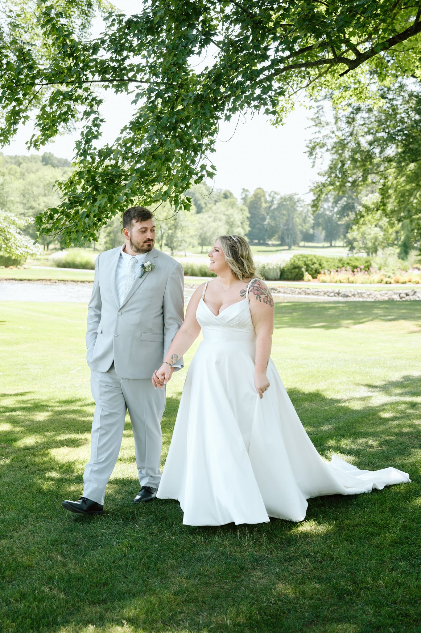 Bride and groom hold hands while walking on green grass beneath a leafy tree on their wedding day.