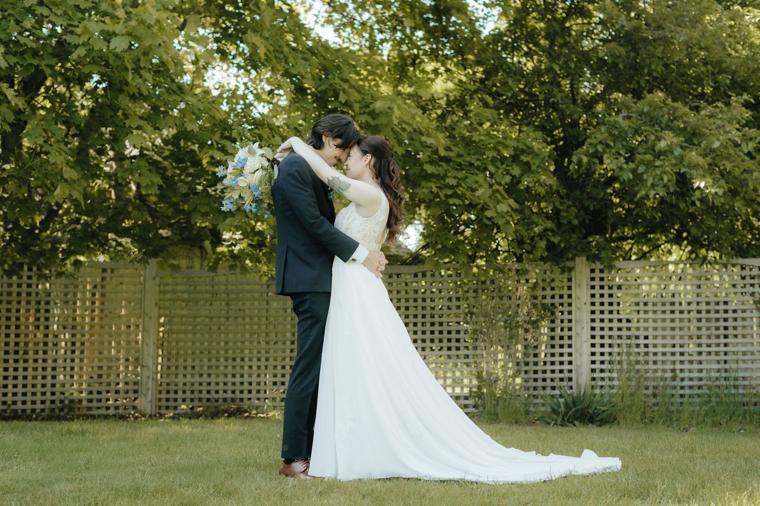 Bride and groom sharing a romantic kiss outdoors on green grass with lush trees and a lattice fence backdrop.