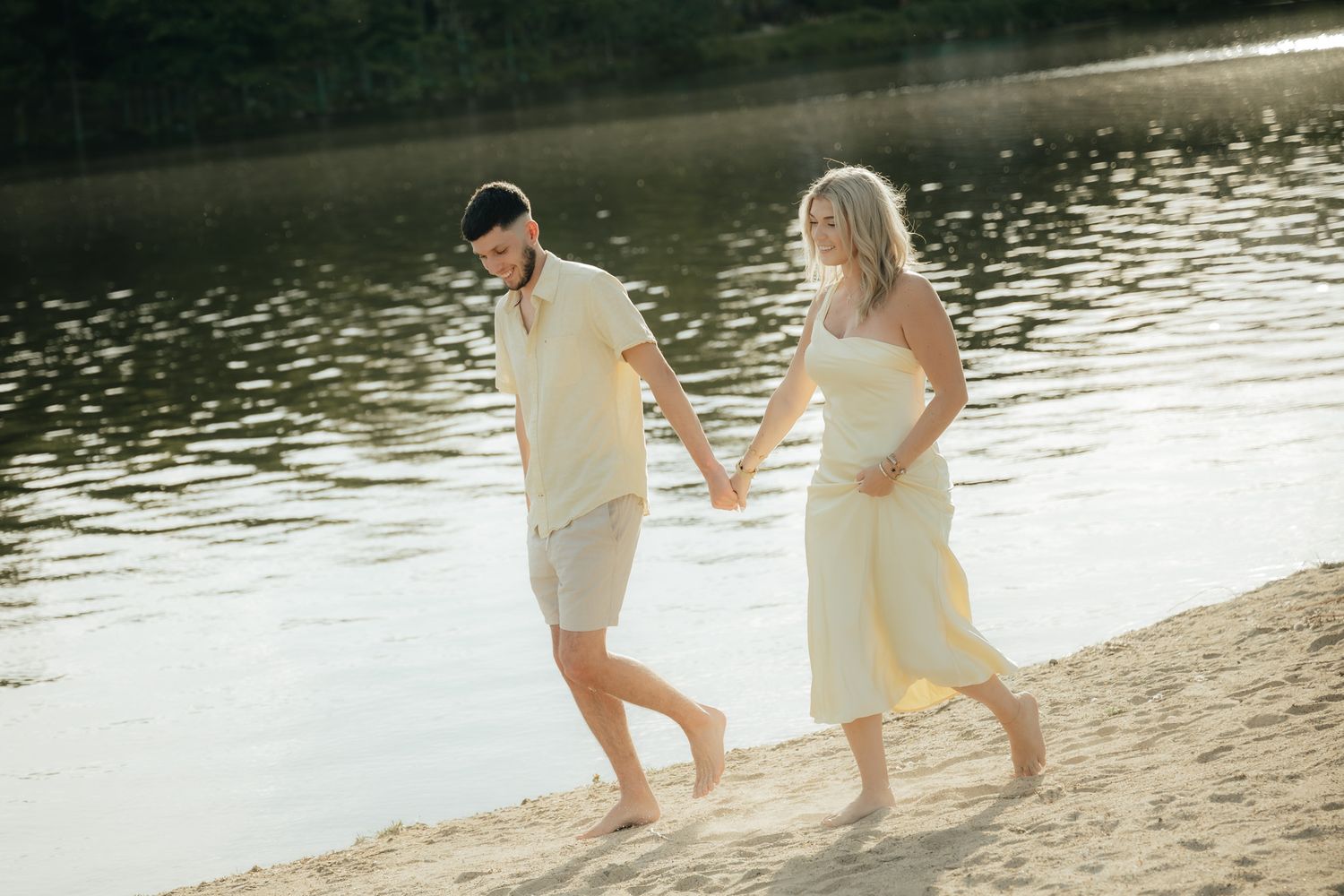 Couple holding hands walking barefoot on sandy beach by calm lake in warm golden sunlight.
