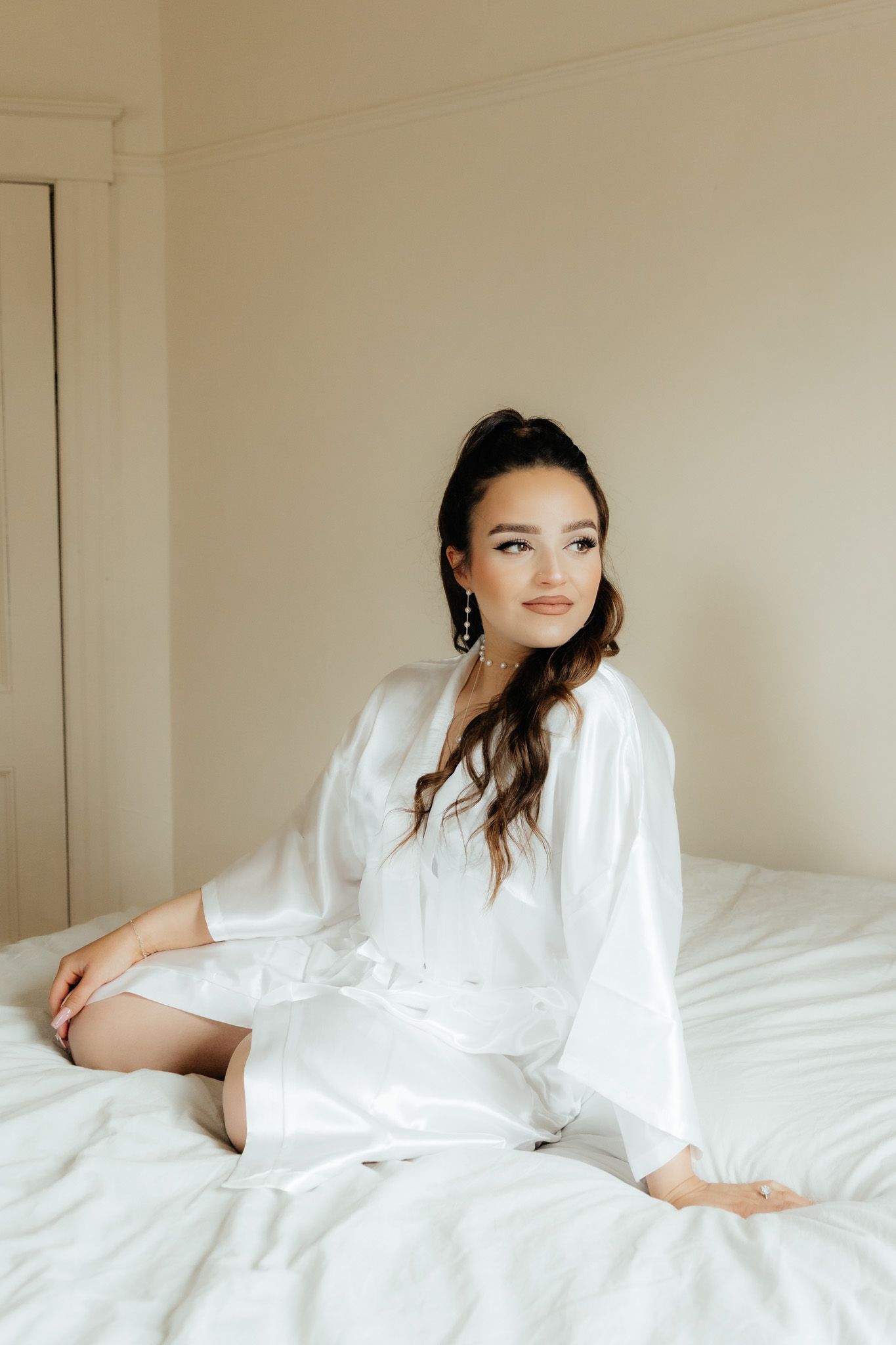 Brunette bride in white silk robe sits gracefully on a white bed.