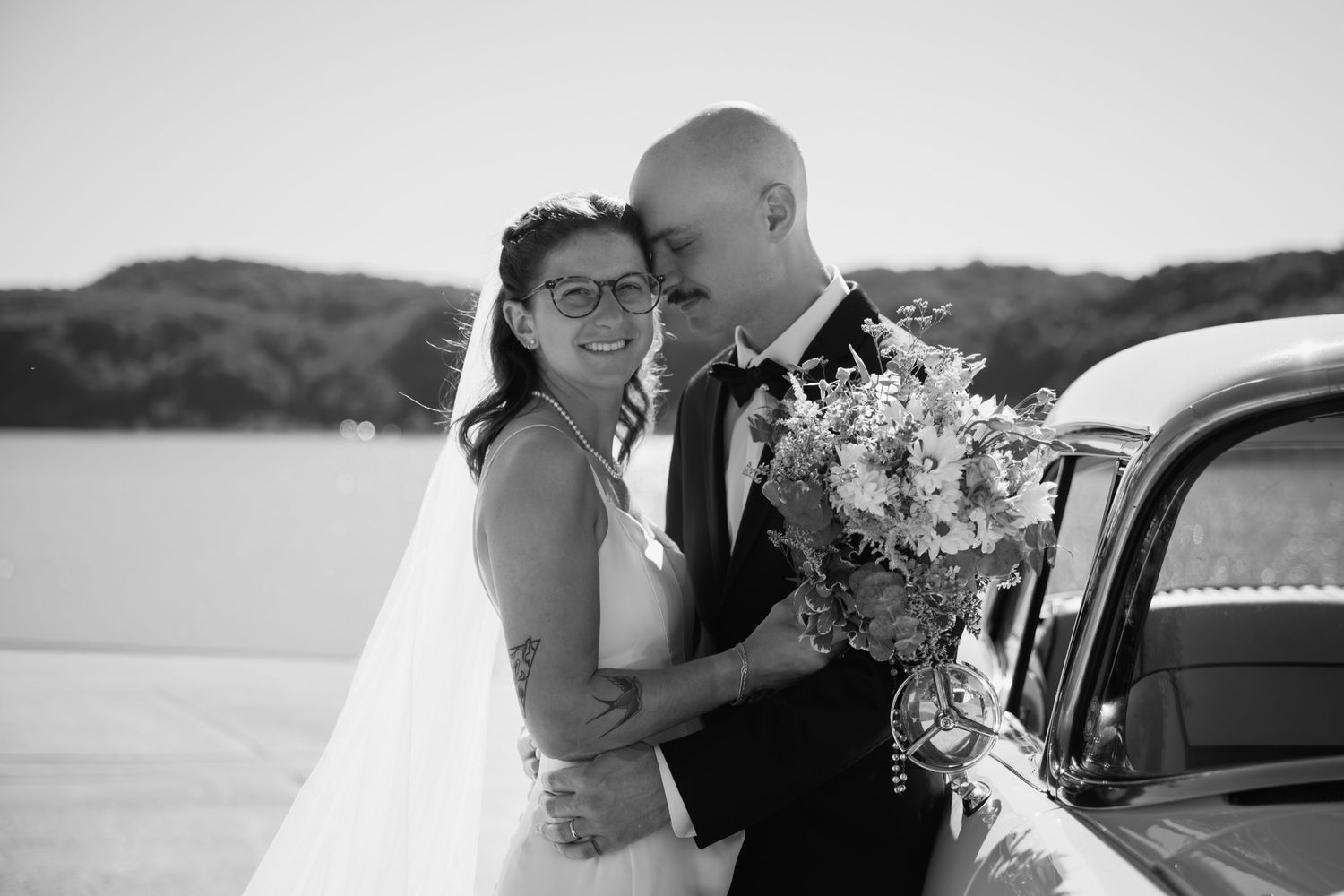 Black and white photo of newlyweds embracing near a vintage car with a lake backdrop.